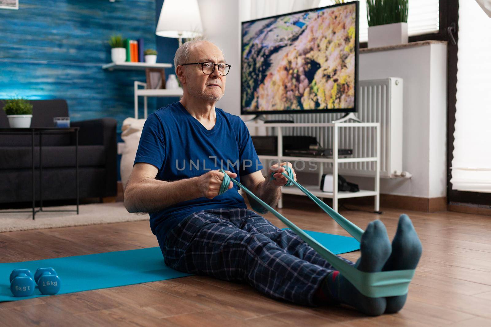 Retirement senior man sitting on yoga mat stretching legs muscles using resistance elastic band training body flexibility. Pensioner in sportswear slimming weight during muscle training in living room