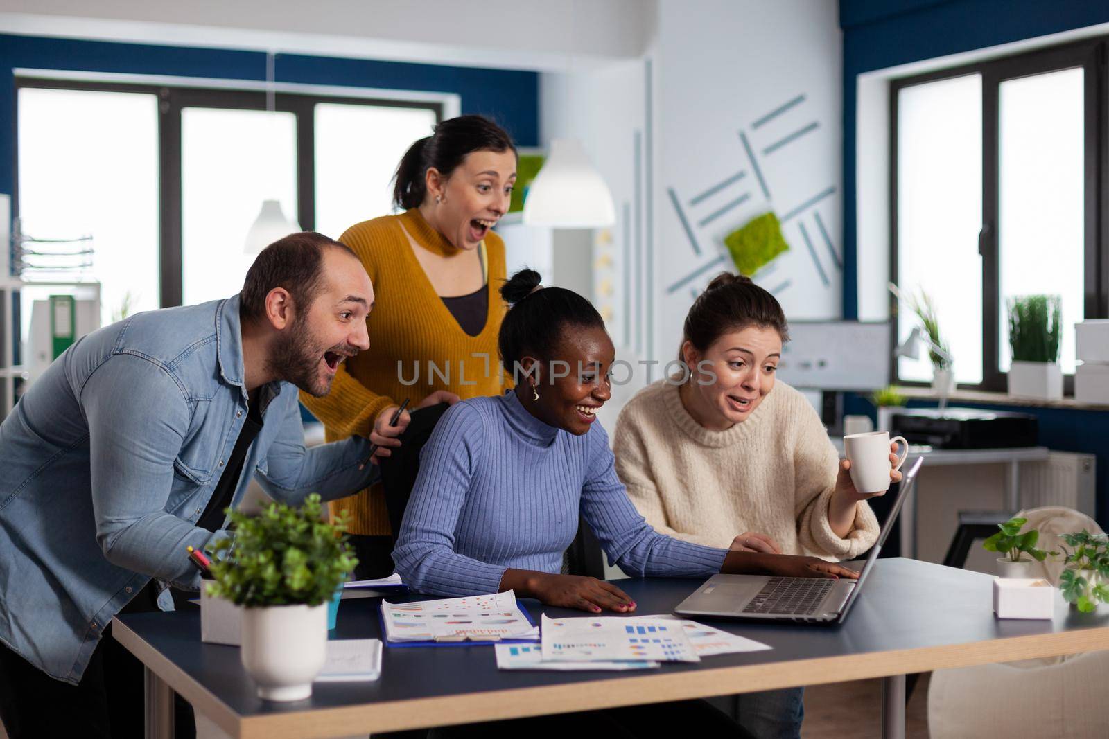 Joyful executive businessman and colleagues screaming in startup after client triumph. Multiethnic diverse positive business team with laptop and papers excited about project.