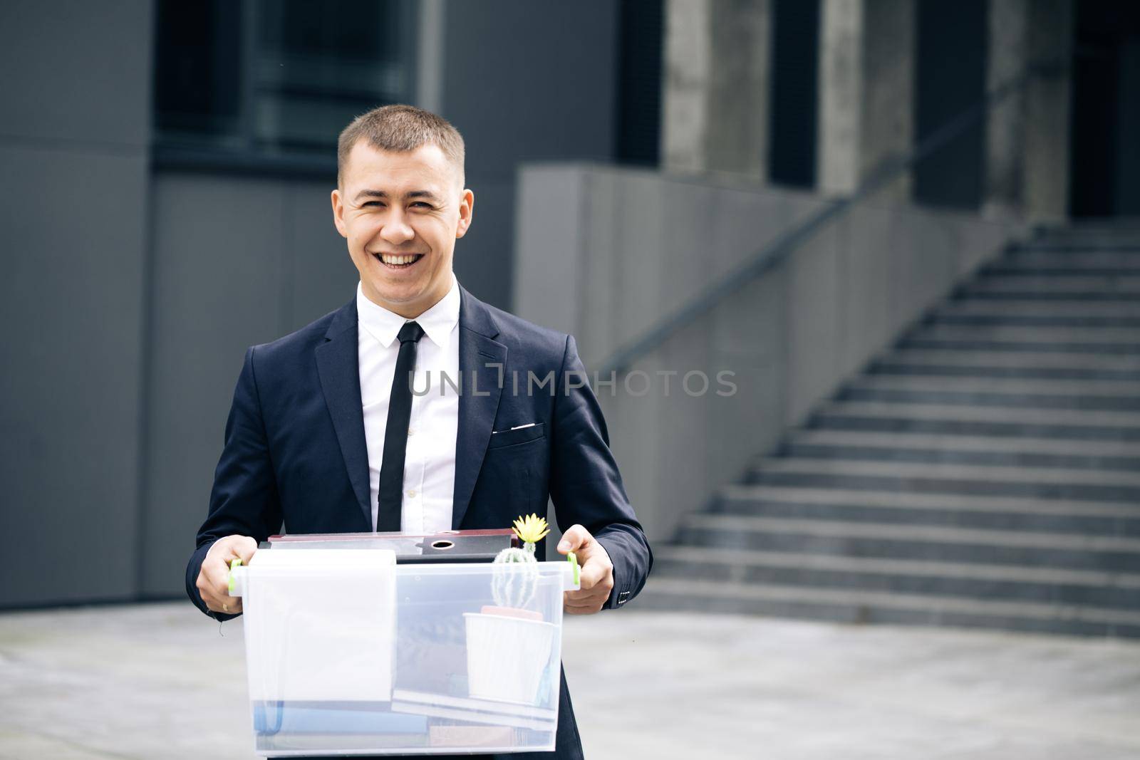 Portrait happy male office worker with box of personal stuff. Unemployment concept. Left without money. Businessman lost job. Fired man outdoors. Happy Jobless person by uflypro