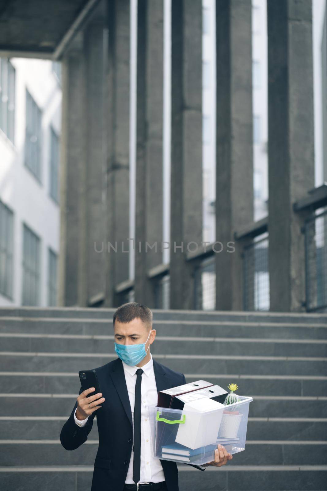 Businessman in medical mask with box of personal stuff walking the street use phone. Business style suit. Coronavirus outdoors social distancing. Finance and industry. Fired man lost job.