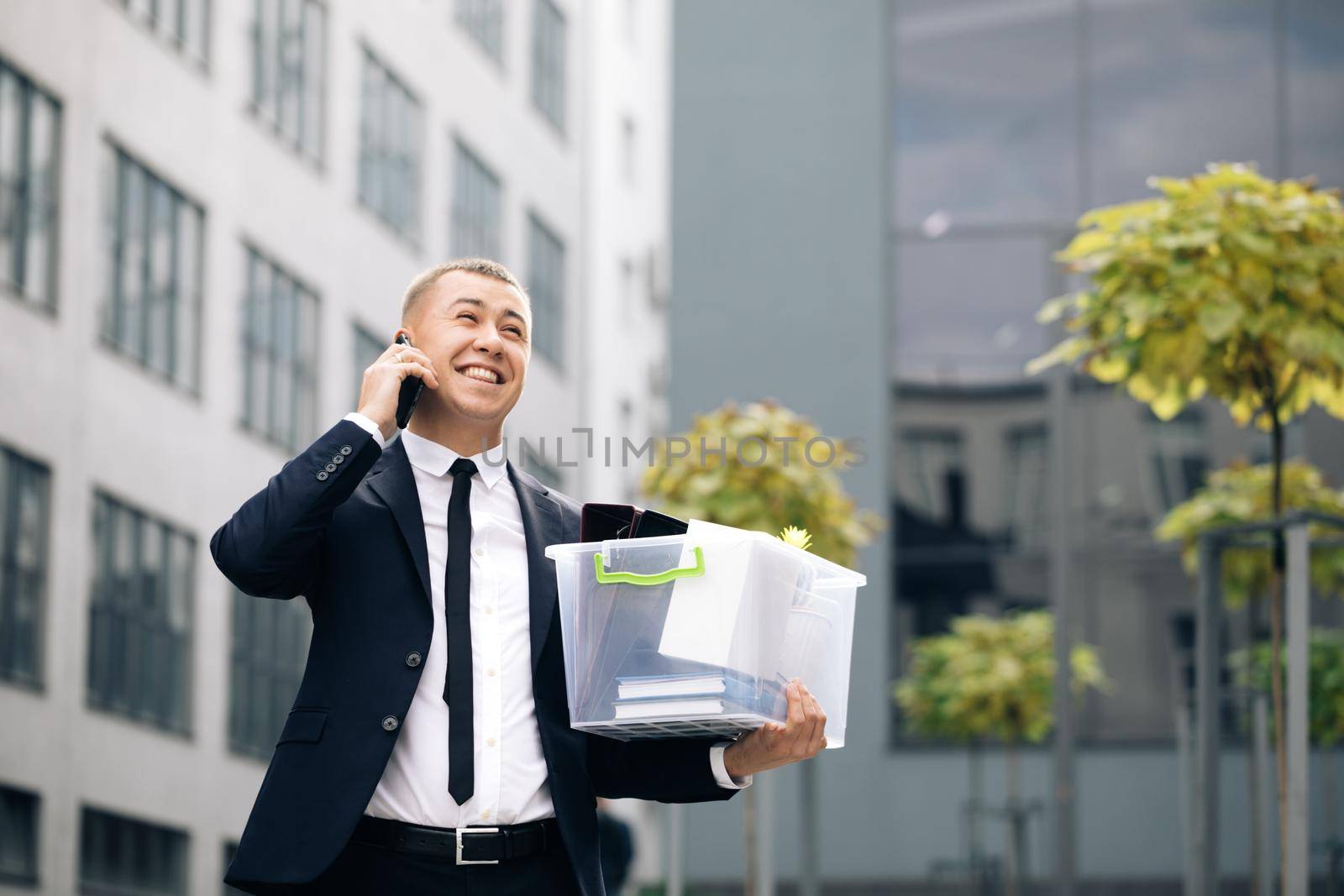 Happy man talking on phone with stuff in box. Cellphone conversation. Speaking on telephone. An employee between office buildings with a box and documents with a desk flower. by uflypro