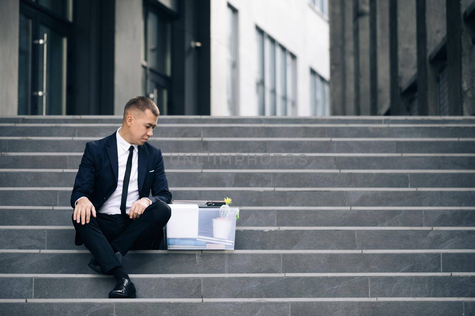 Male office worker in despair lost job. Sad businessman sitting on stairs outdoor with box of stuff as lost business. Fired man. Unemployment rate growing due pandemic by uflypro