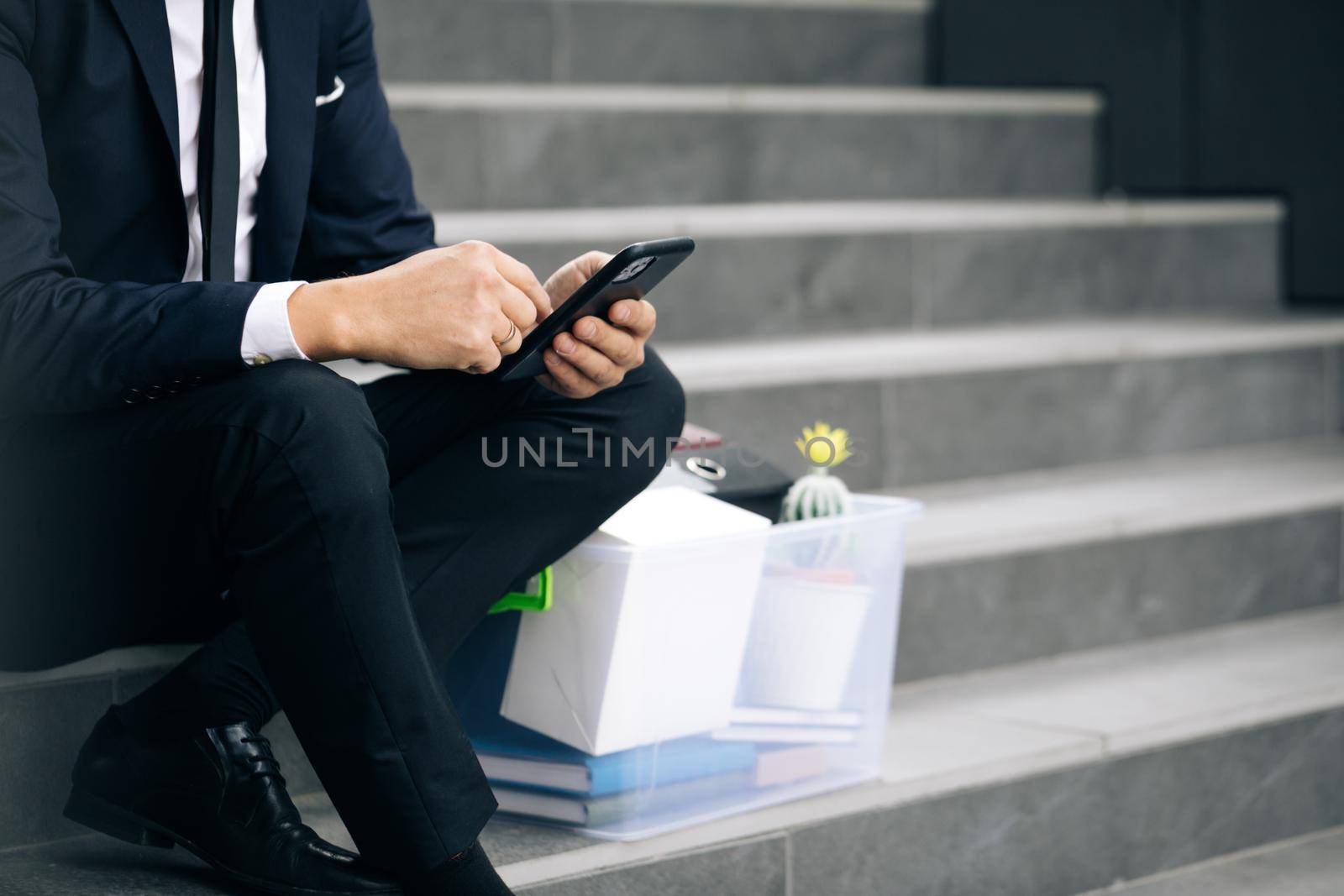 Sad businessman sitting on stairs outdoor with box of personal stuff. Despaired male lost work place and looking for a job on smartphone. Pandemic economical crisis. Fired man by uflypro