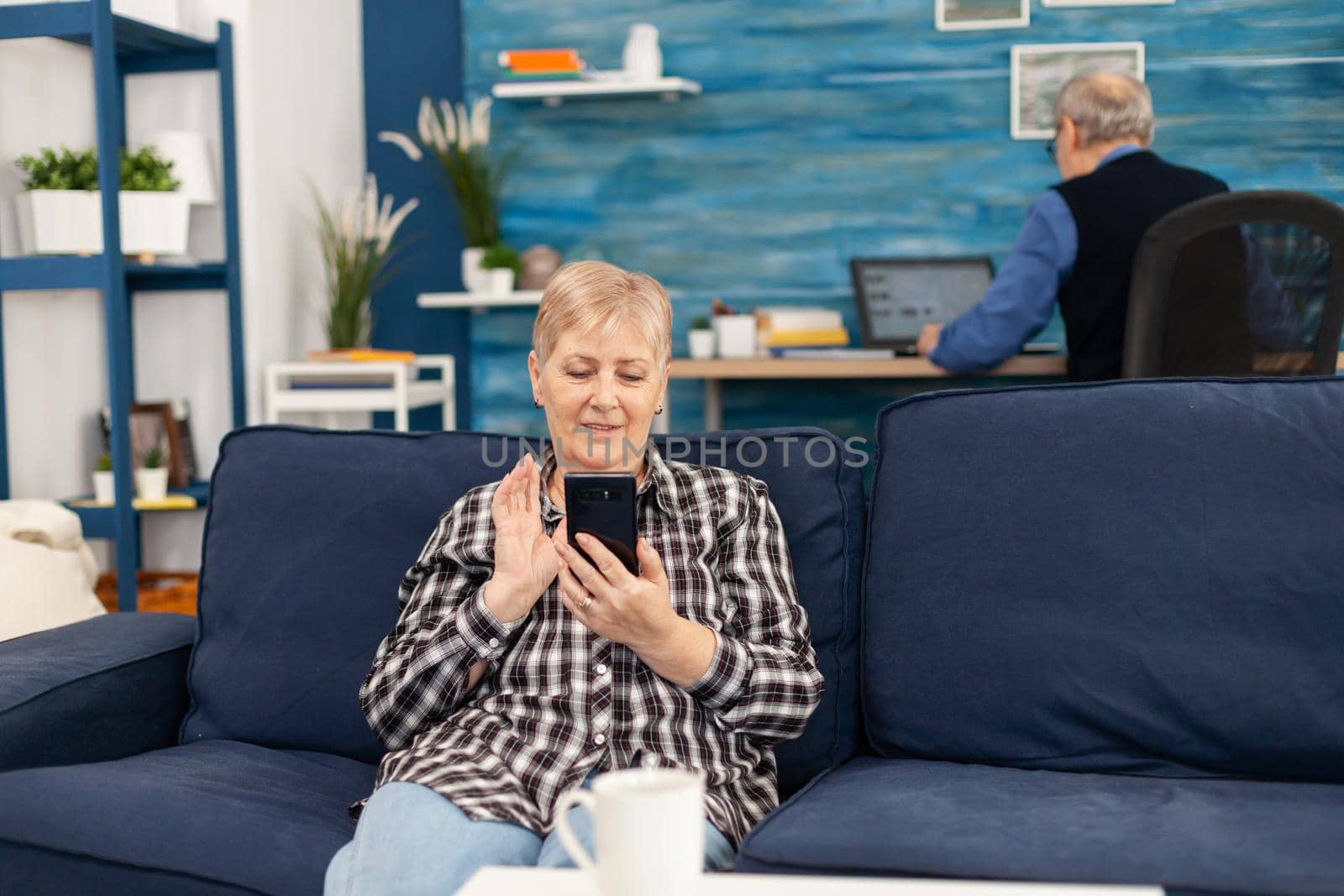 Happy elderly lady waving at smartphone during video call. Senior woman waving at phone webcam in the course of video conference sitting on living room sofa.