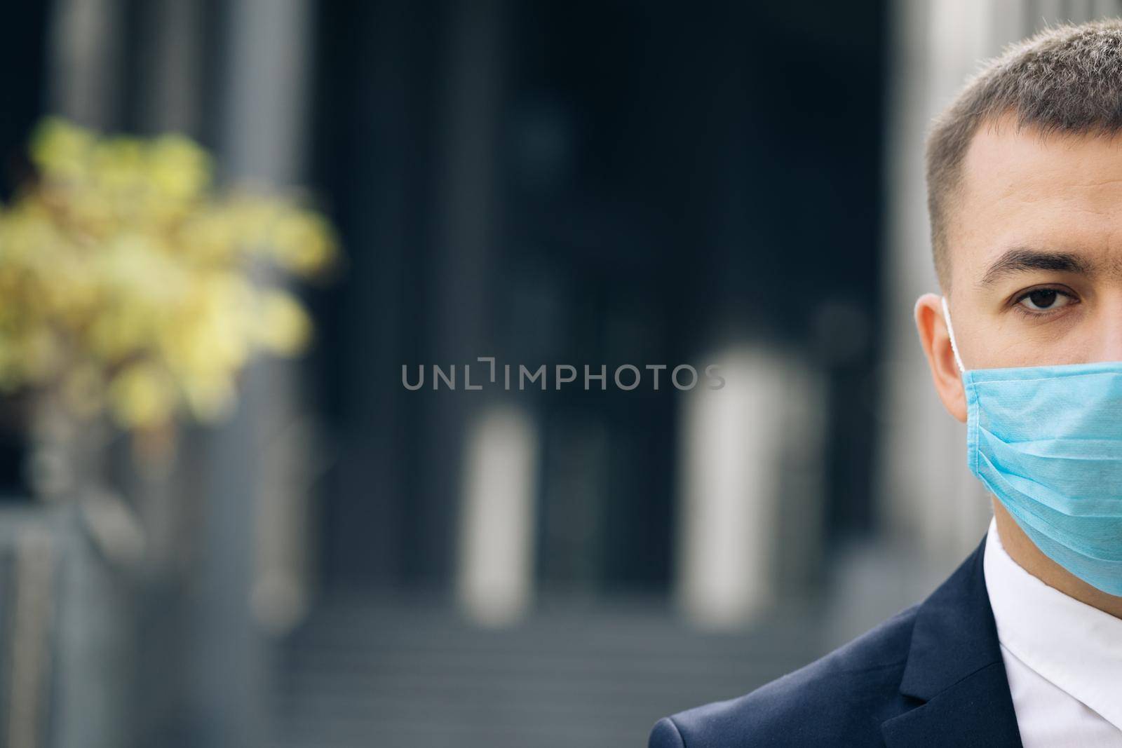 Close up of half face of upset caucasian young handsome man in medical mask looking straight to camera while standing outdoors near modern office building. Man in protection mask look at street.