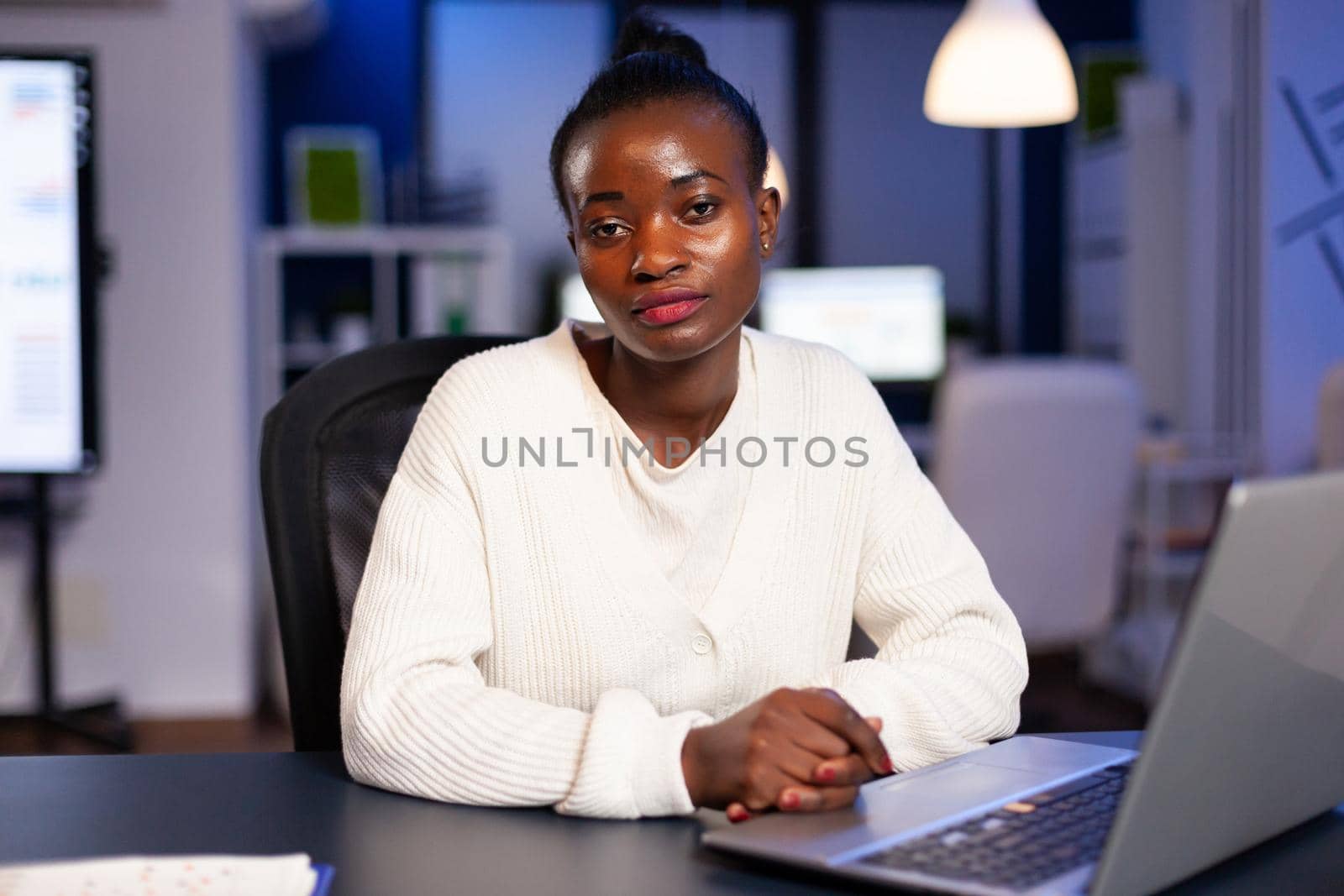 Focused tired business woman looking at camera after reading tasks on laptop sittting at desk in start-up company late at night. Employee doing overtime for job respecting deadline