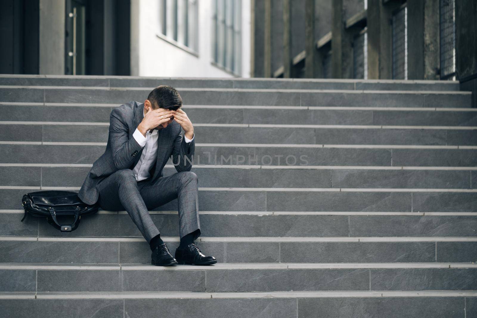 Fired unhappy european bearded man losing job sitting on stairs. Upset young businessman in frustration outside office department. Unemployment. Crisis by uflypro