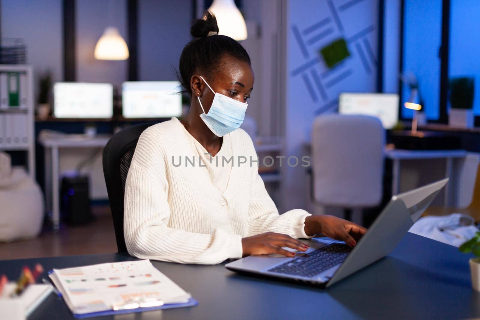 African woman with face mask reading emails late at night to respect deadline of project working in new normal business office, analysing documents, making strategy overtime during global pandemic.