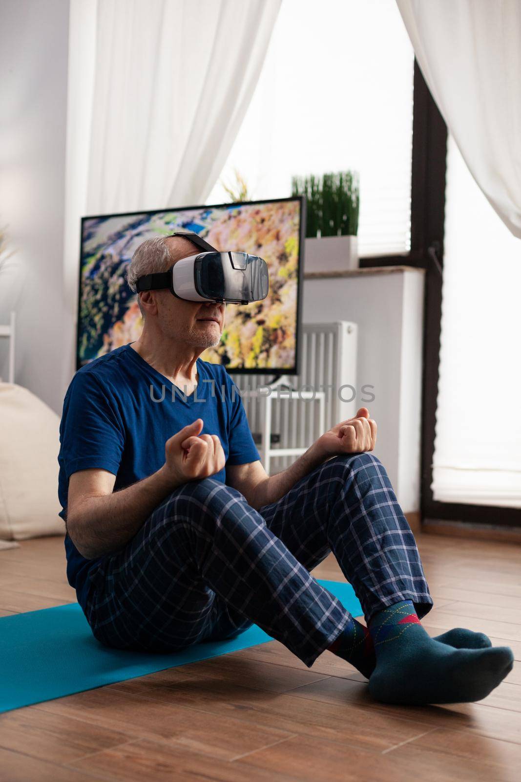 Zen senior man sitting on yoga mat in lotus position during pilates workout in living room stretching body muscle. Pensioner wearing virtual reality headset exercising meditation