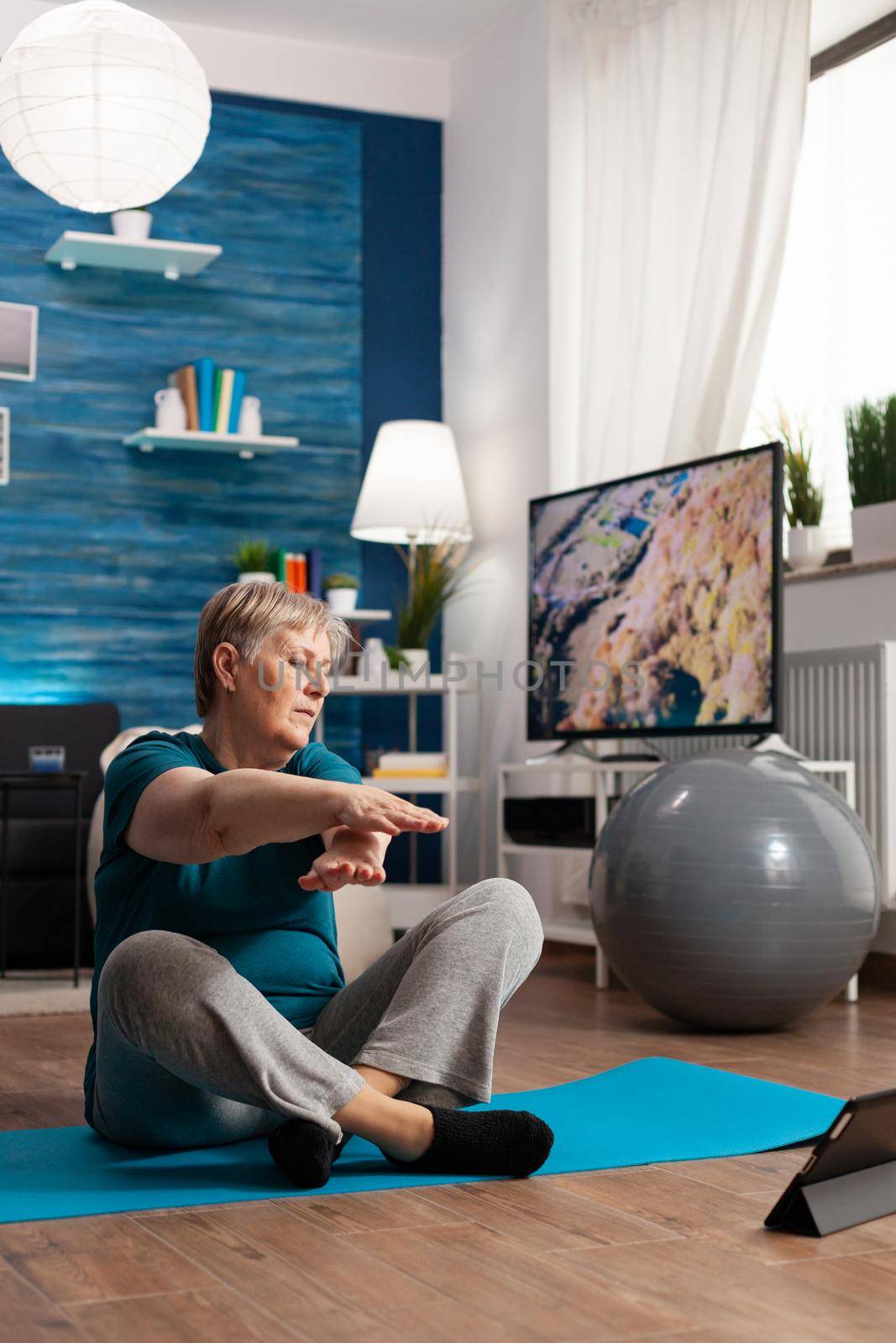 Retired senior woman in sportswear sitting on yoga mat watching fitness lesson on laptop practicing arm exercise slimming weight. Pensioner doing working at body flexibility during wellness workout