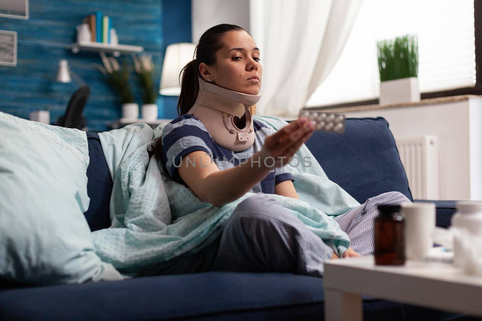 Woman in neck brace suffering from pain on sofa after bad accident injury. Young caucasian adult with ache taking medicine for back complication, physical muscular contracture