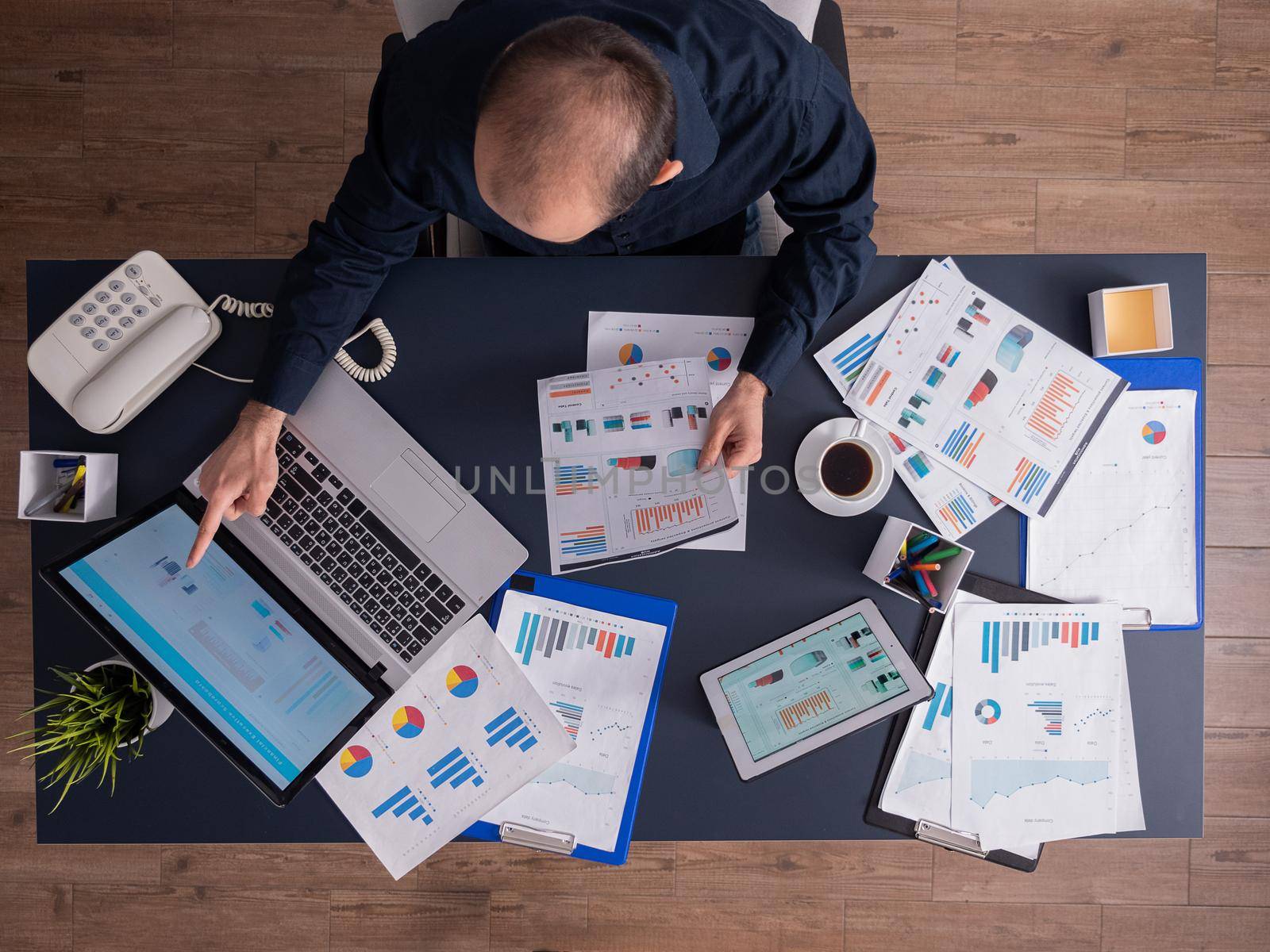 Top view of businessman pointing at financial statistics on laptop monitor sitting at desk in corporate office with papperwork. Employee using tablet pc with charts.