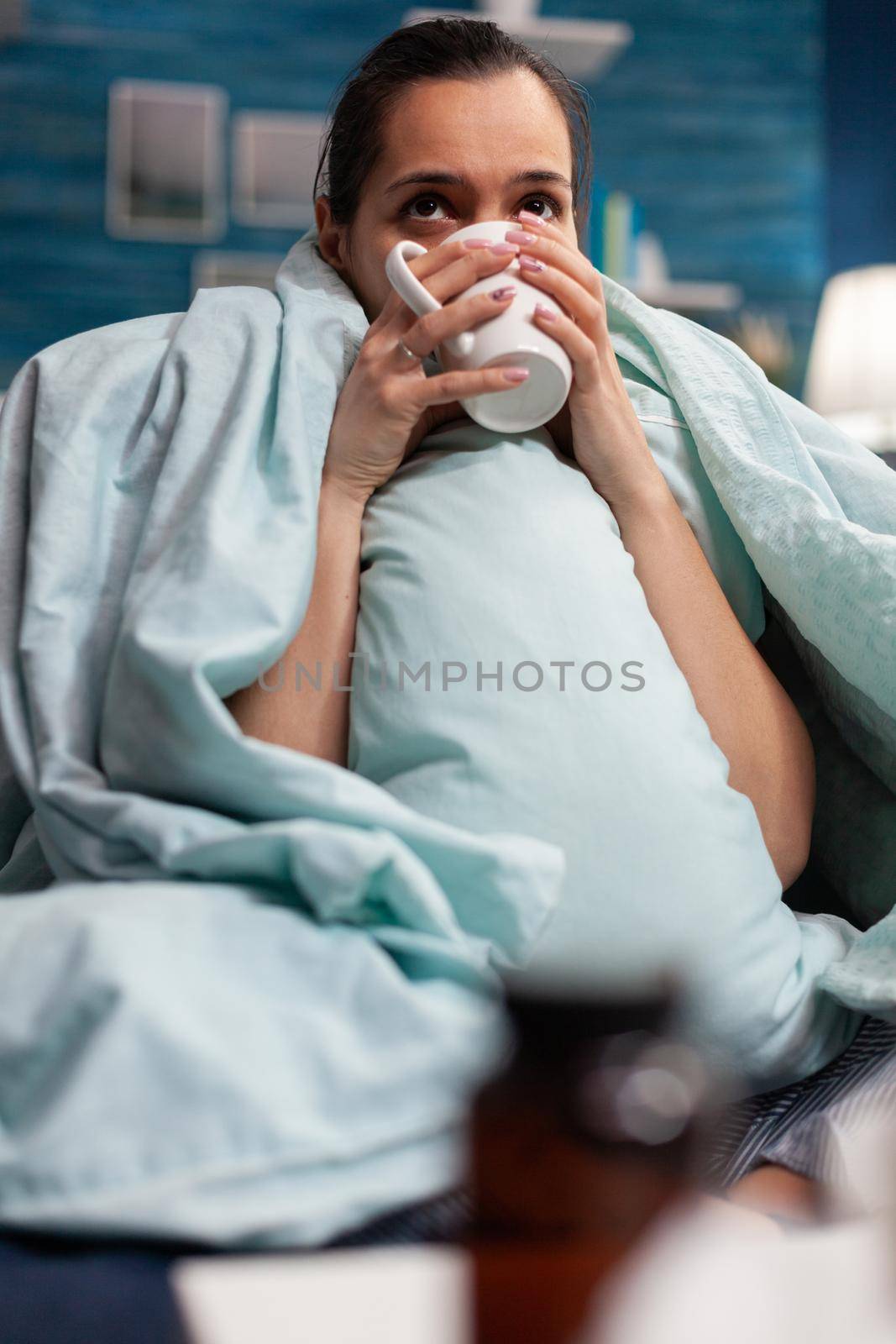 Sick woman at home resting with cup of tea on sofa wrapped in blanket. Unwell young adult with cold flu disease infection fever temperature headache covid 19 virus symptoms