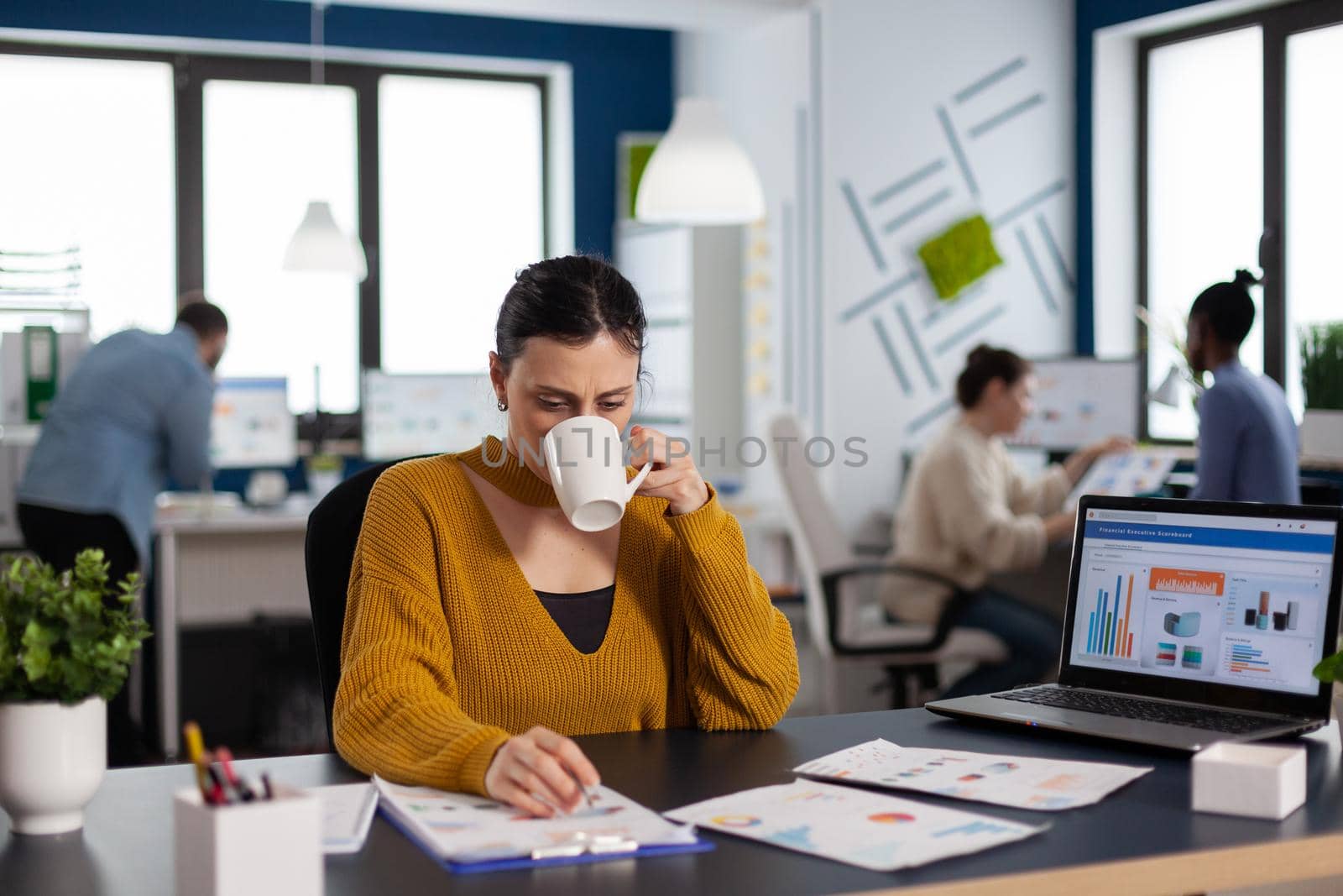 Brand manager businesswoman drinking a cup of coffee analysing financial start up charts. Executive entrepreneur, manager leader sitting working on projects with diverse teamworkers.