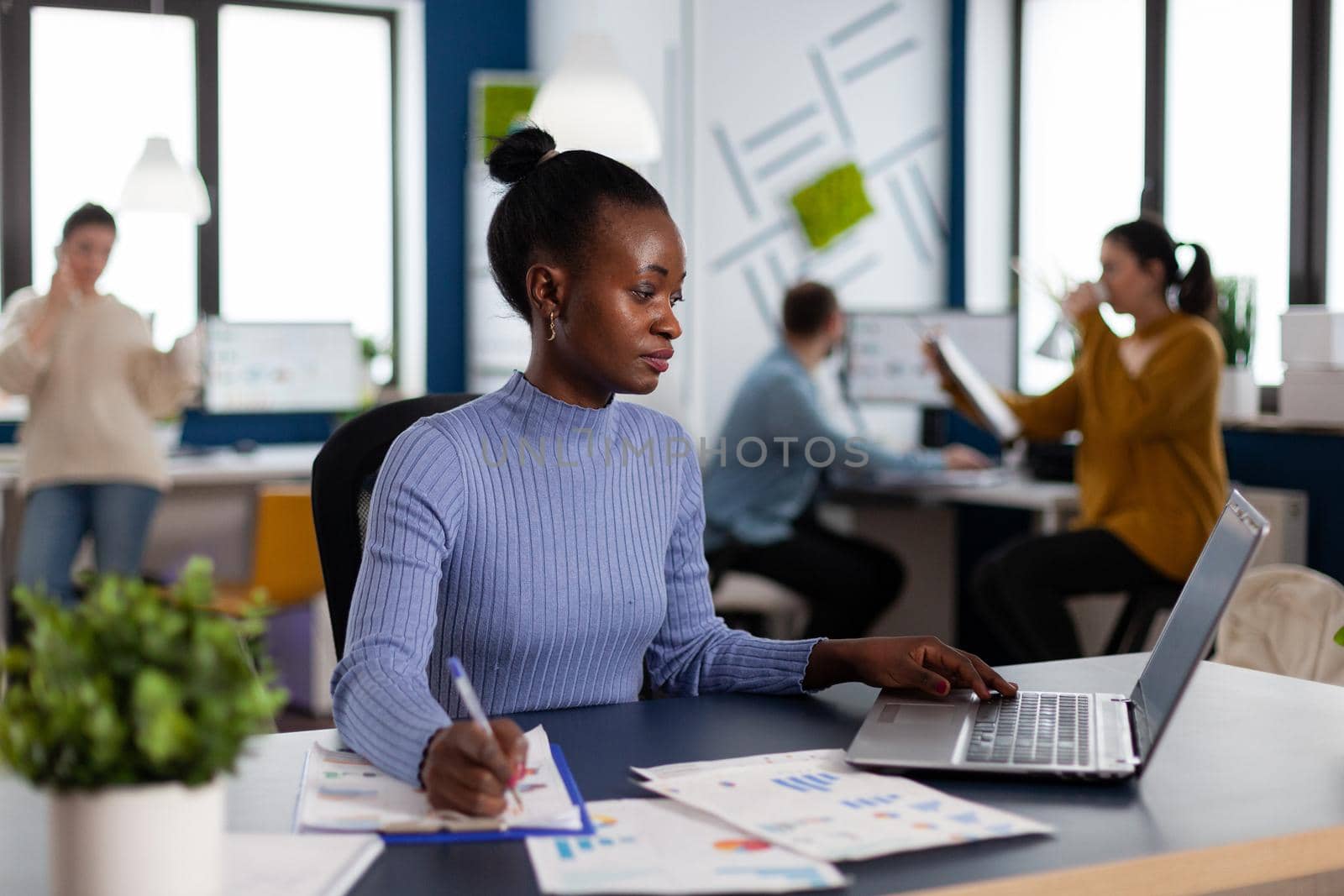 African businesswoman taking notes on clipboard analysing statistics in start up office. Diverse team of business people analyzing company financial reports from computer. African businesswoman taking notes on clipboard analysing statistics
