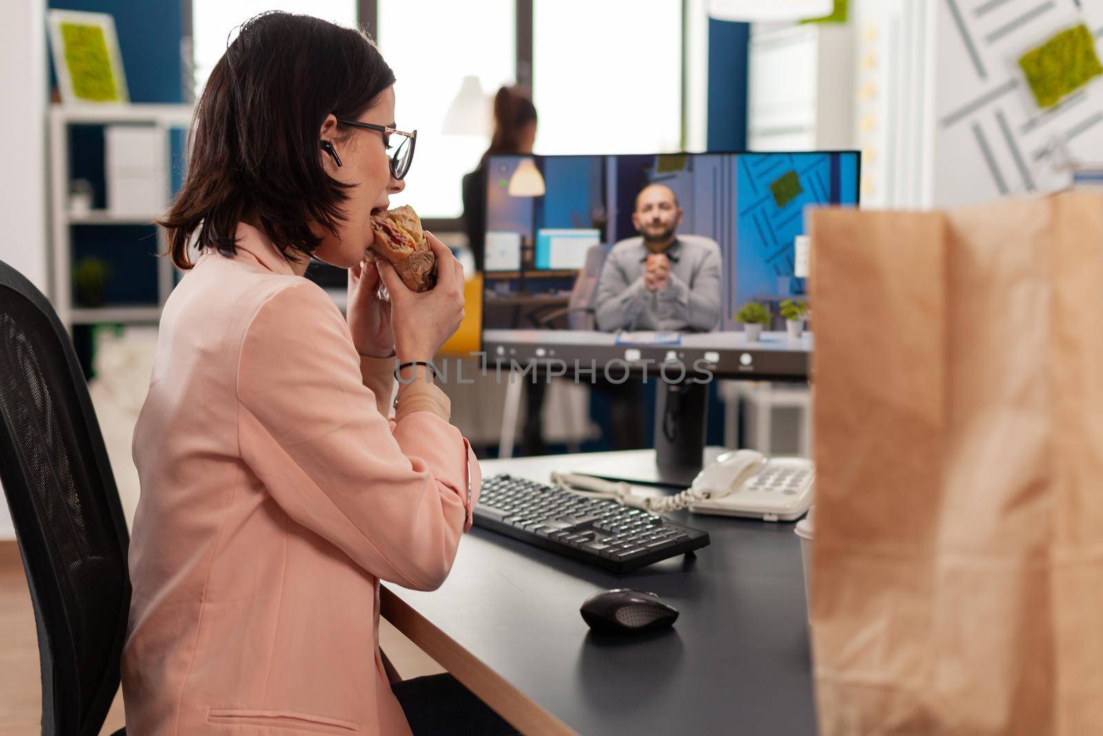 Businesswoman eating delivery takeaway sandwich during online videocall conference by DCStudio