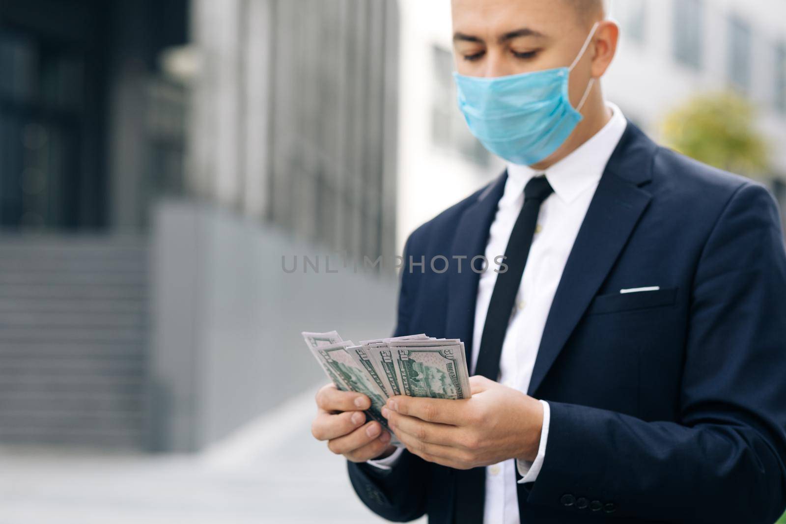 Happy good looking man in business style holding dollars and calculating. Cheerful rich handsome businessman in protective face mask counting money near business center background.