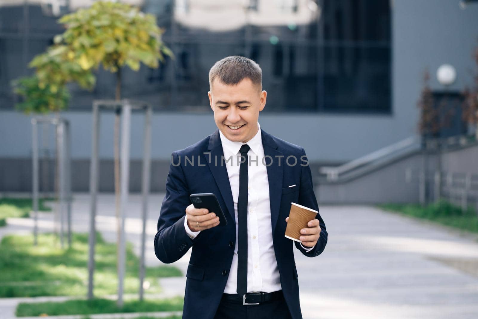Stylish Businessman Exiting from the Modern Office. Using his Smartphone. Sliding on Mobiles Screen. Drinking Tasty Coffee. Looking Successful, Confident. Holding Coffee Cup on Summer Day.