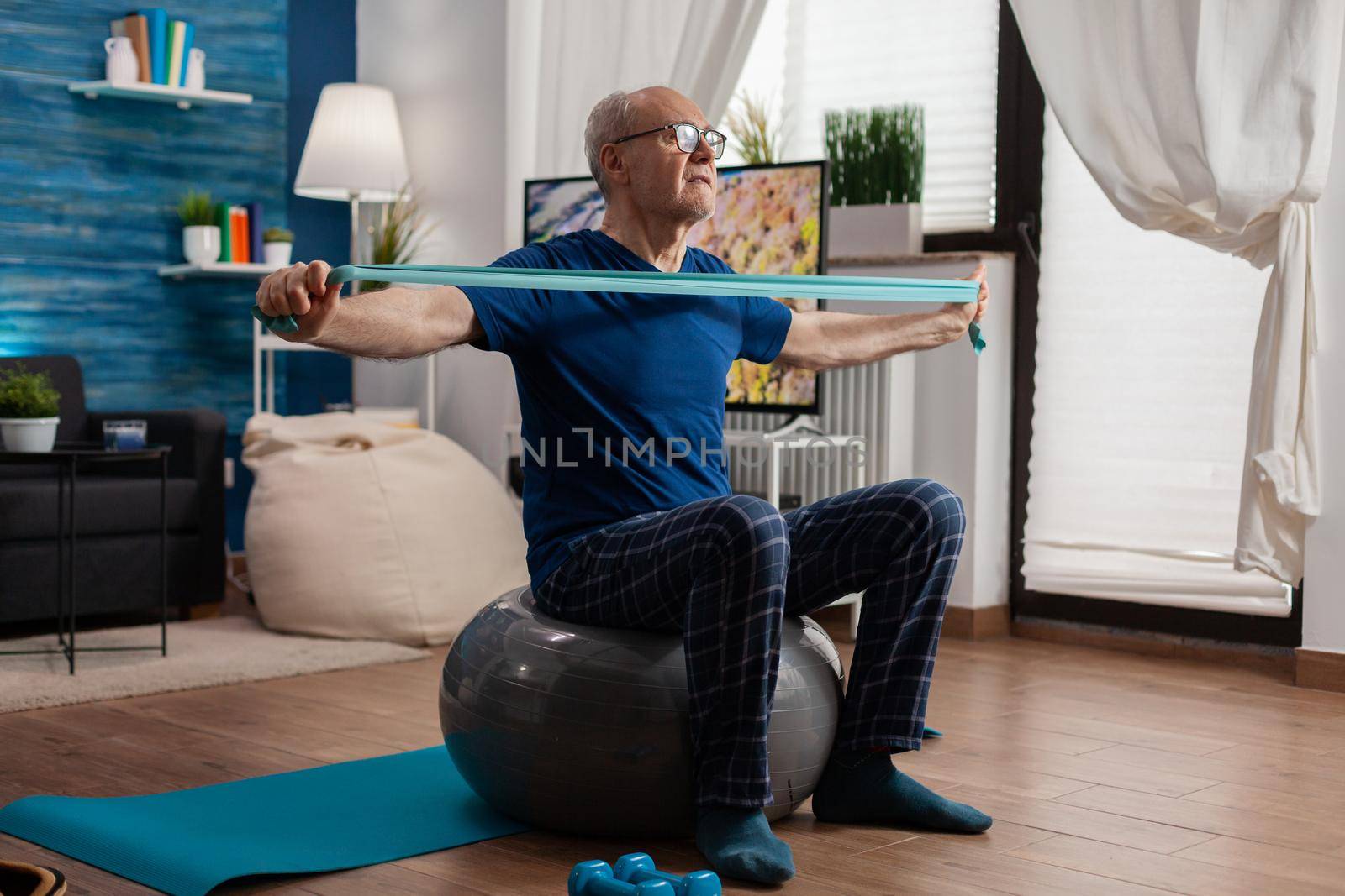 Retiree senior man sitting on swiss ball in living room doing healthcare fitness exercises streching arm muscles using resistance elastic band. Pensioner training body strength in living room