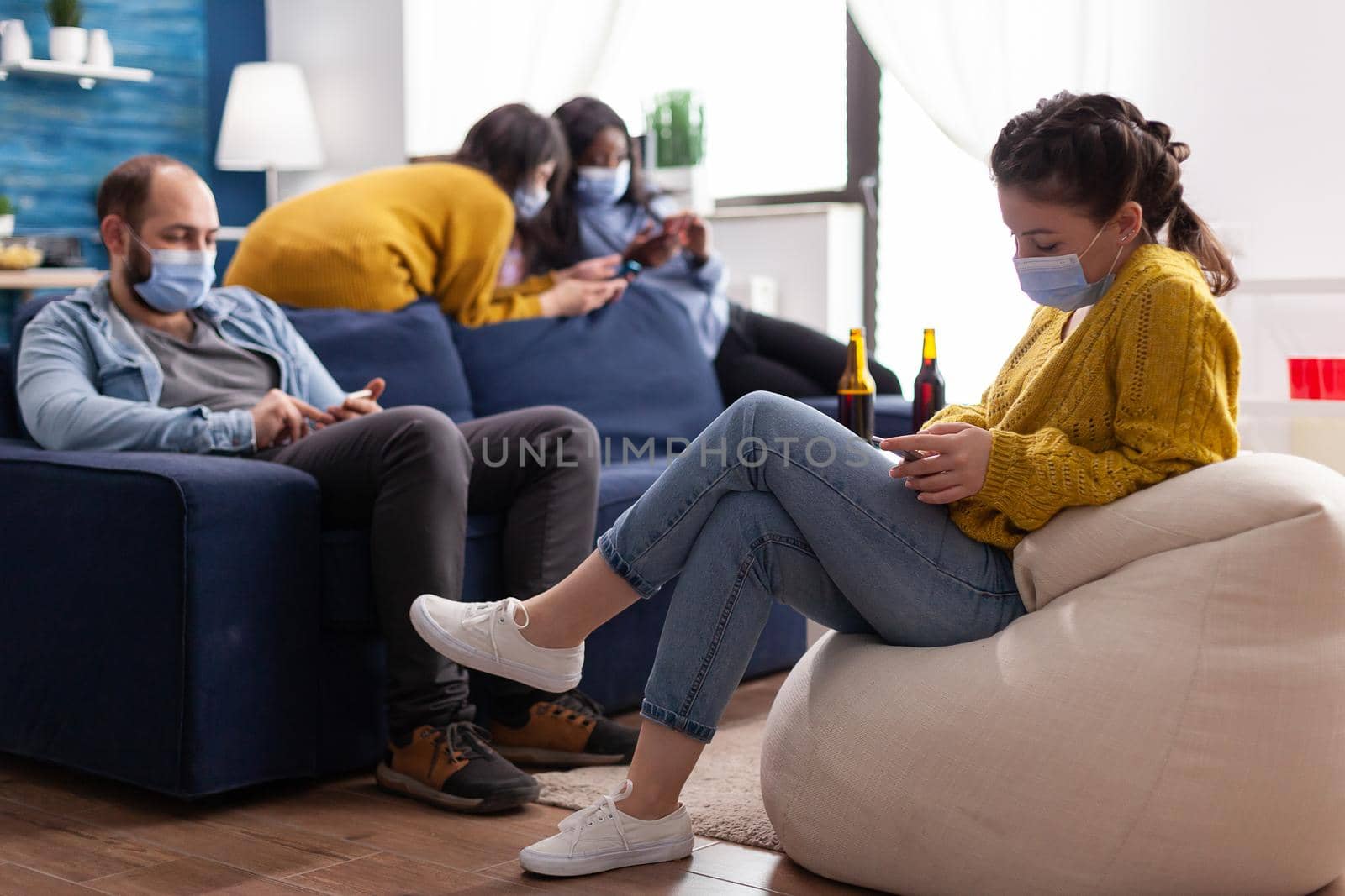 Group of mixed race friends browsing on smartphone keeping social distancing wearing face mask to prevent spread of coronavirus during global pandemic in living room with beer bottle. Conceptual image