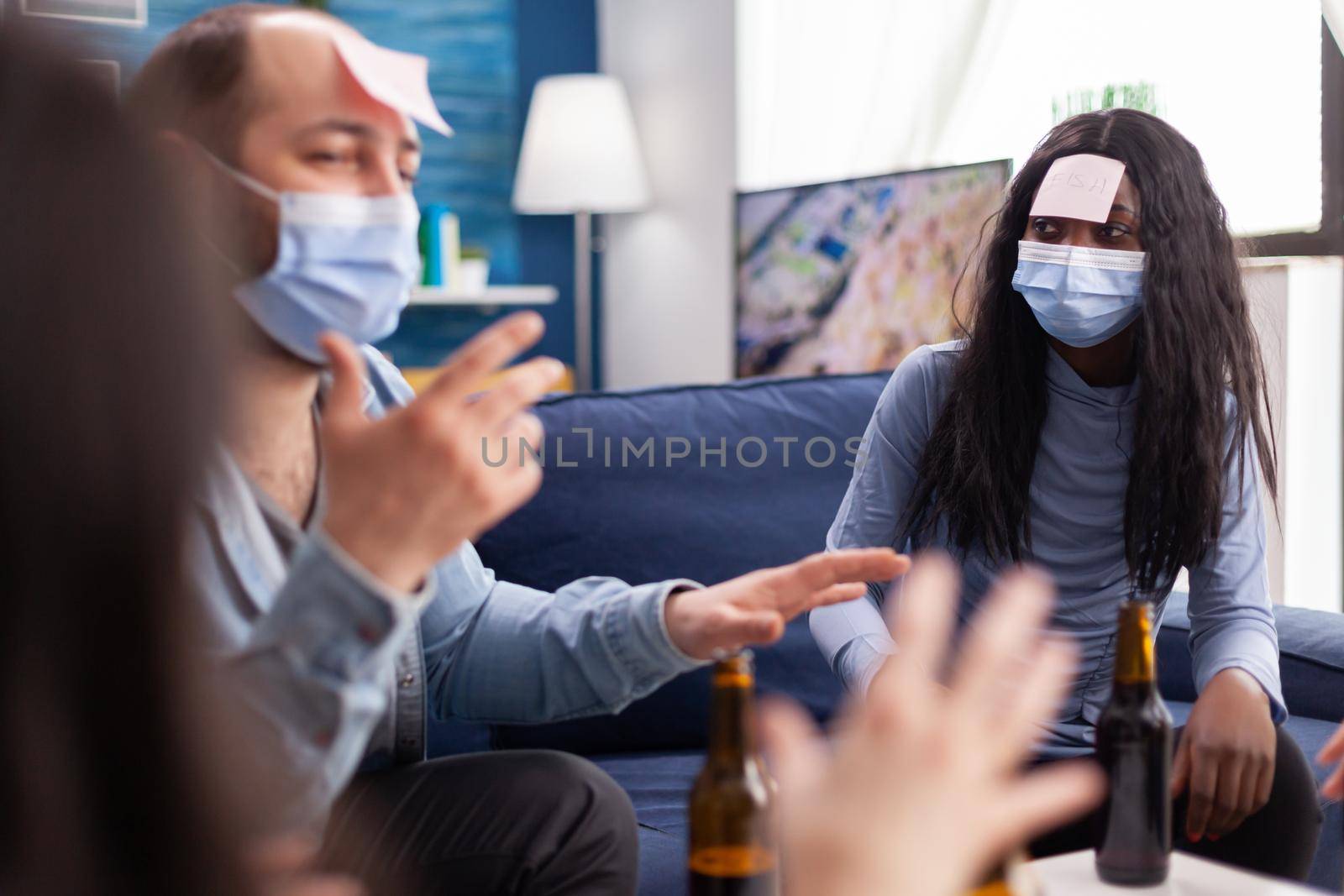 African woman with sticky note on forehead playing name game with cheerful happy friends wearing face mask to prevent infection with covid19 due to social pandemic. Beer bottle,