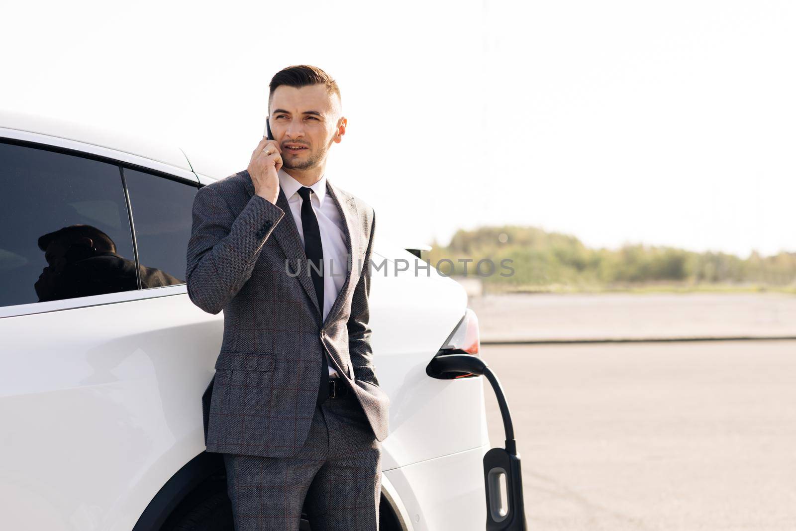 Portrait of handsome joyful 30-aged businessman with neat beard which has mobile conversation while own luxurious car charging battery on specially-equipped charging station.