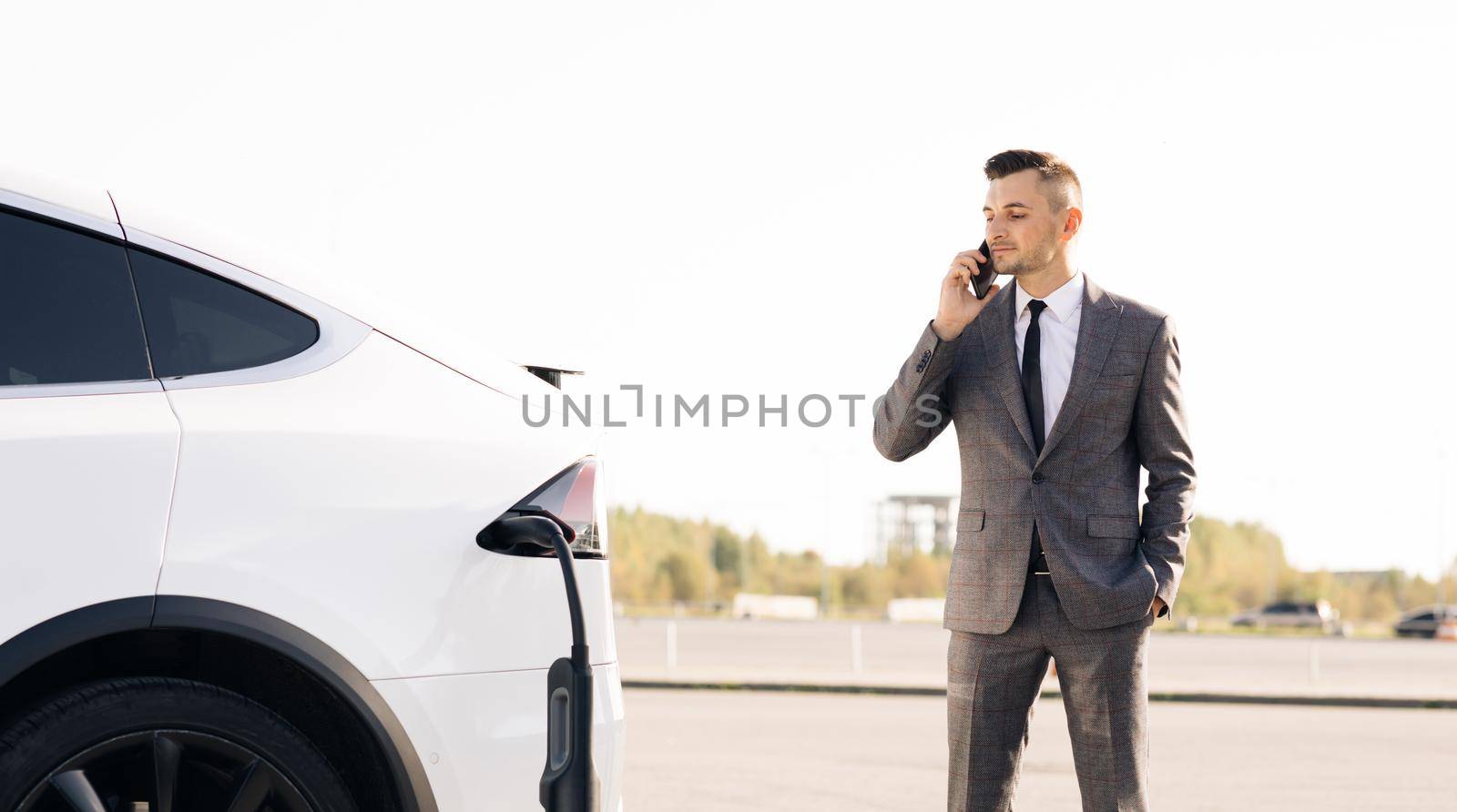 Businessman using smartphone and leans on white luxurious electric car that is charging. Plug charging an electrical car. Luxury electrical car recharging.