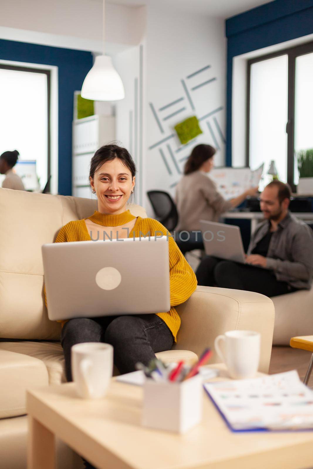 Young woman entrepreneur sitting on couch in middle of business start up office looking at camera smiling holding laptop. Multiethnic coworkers talking about startup financial company in modern office