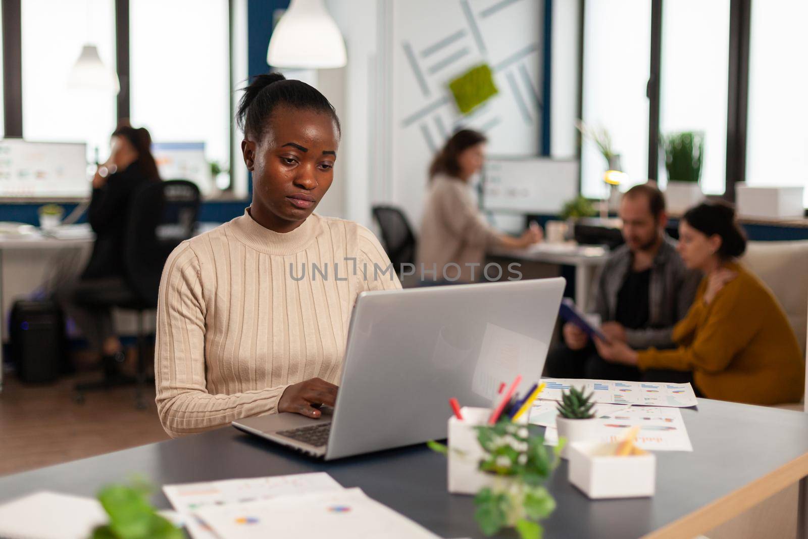 Portrait of authentic african business woman reading email on laptop sitting at desk in busy start up office while diverse team analyzes statistics data. Multiethnic team working at new project