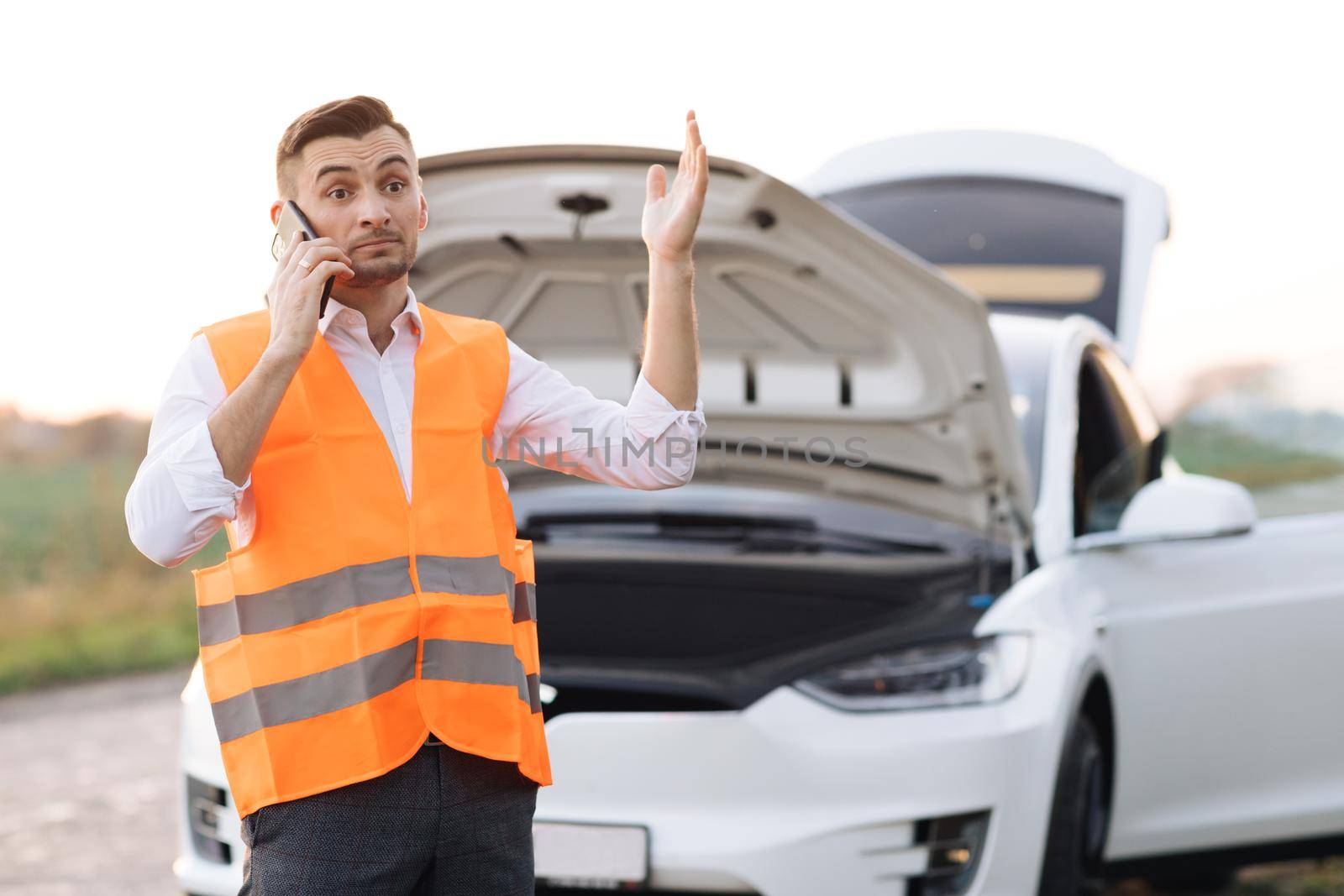 Young man outside car. Dissapointed businessman talking on phone. Broken car with opened hood stand besides. Helpless man need help and assistance. Electric car accident. Car trouble. by uflypro