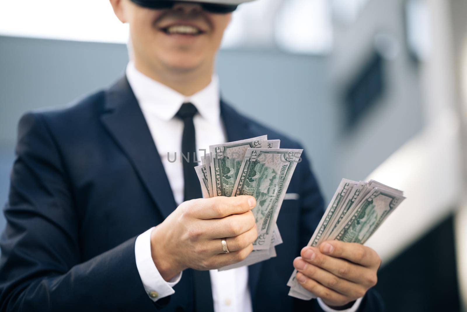 Rich man wearing VR headset counting money and smiling. Young businessman standing with pack of dollars. Richness and success concept. New technology offers new 3D dimensions