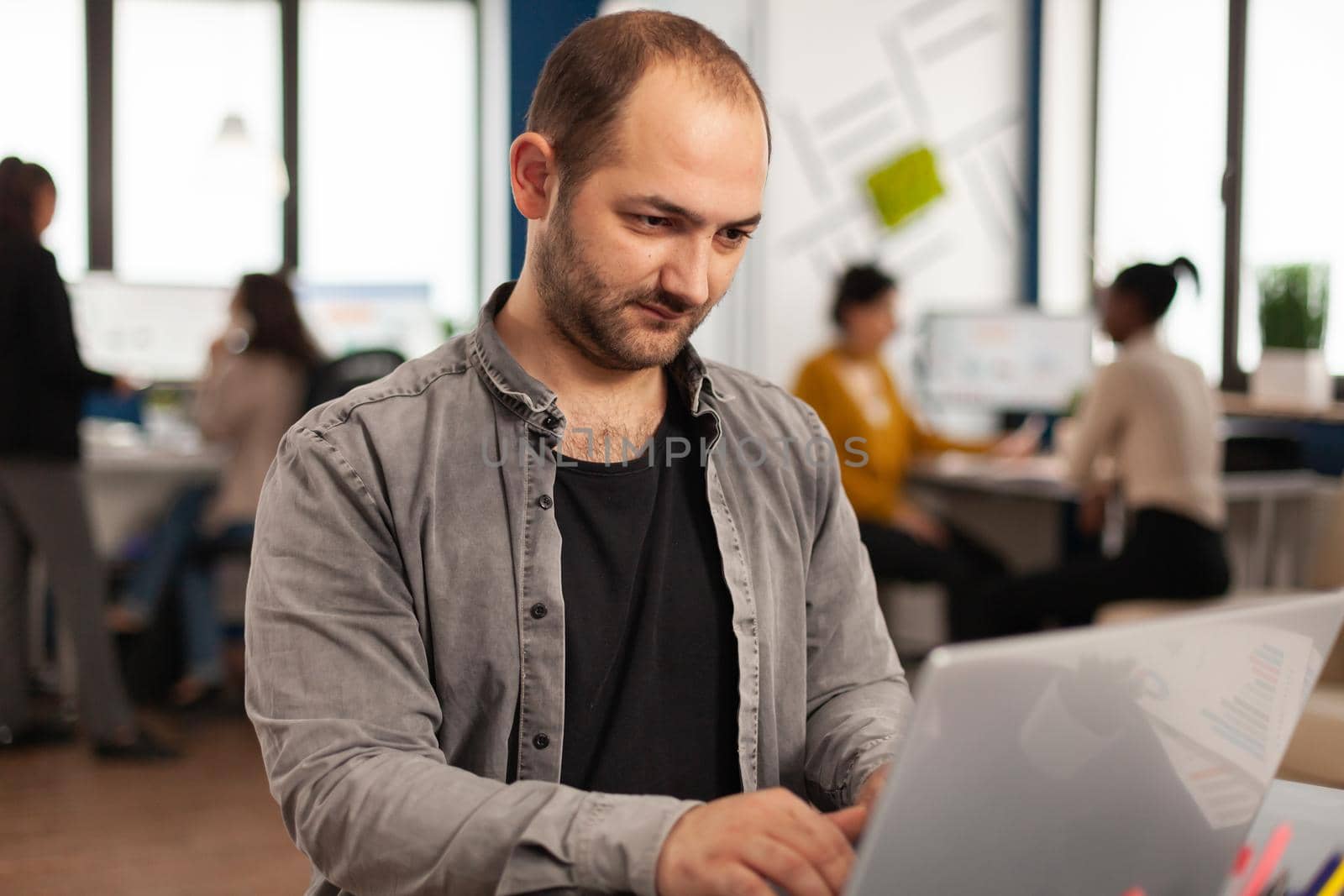 Enthusiastic business man writing a report sitting at table in modern startup business office sending mails typing on laptop, greeting colleague. Man working in financial company with diverse team.