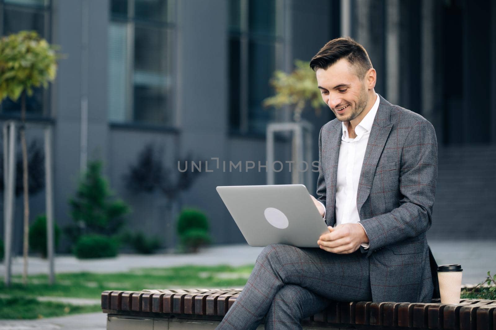 Modern businessman working with laptop on the bench outdoors. Handsome talking video conference on notebook near office. Male professional explaining project with gestures at remote workplace