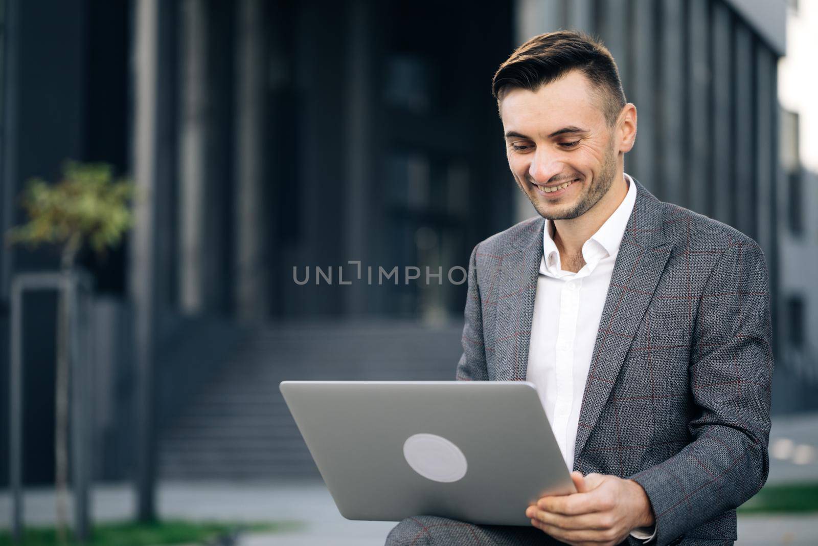 Close up of confident businessman working on laptop outdoors. Stylish successful entrepreneur talking on video conference on laptop. Modern businessman working with laptop on the bench outdoors.