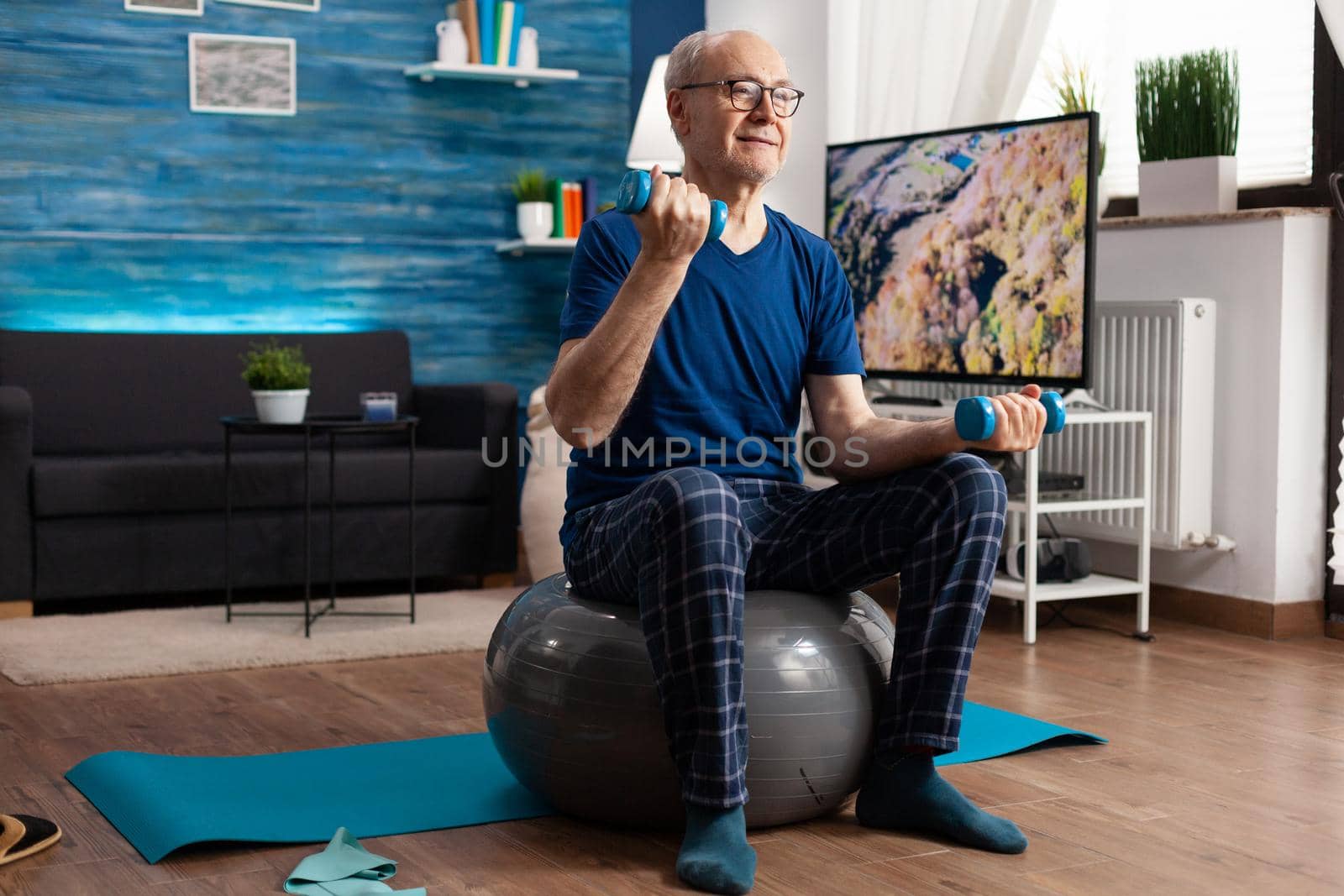 Senior man holding dumbbells doing arms exercise streching body muscles training muscular resistance. Retirement caucasian male sitting on swiss ball in living room doing fitness workout