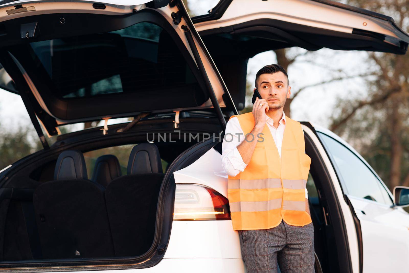 Car accident on the road. Man with reflective vest calling by phone about breakdown in her electric car. Emergency stop sign. Caucasian man talking on phone to call someone to help car service. by uflypro