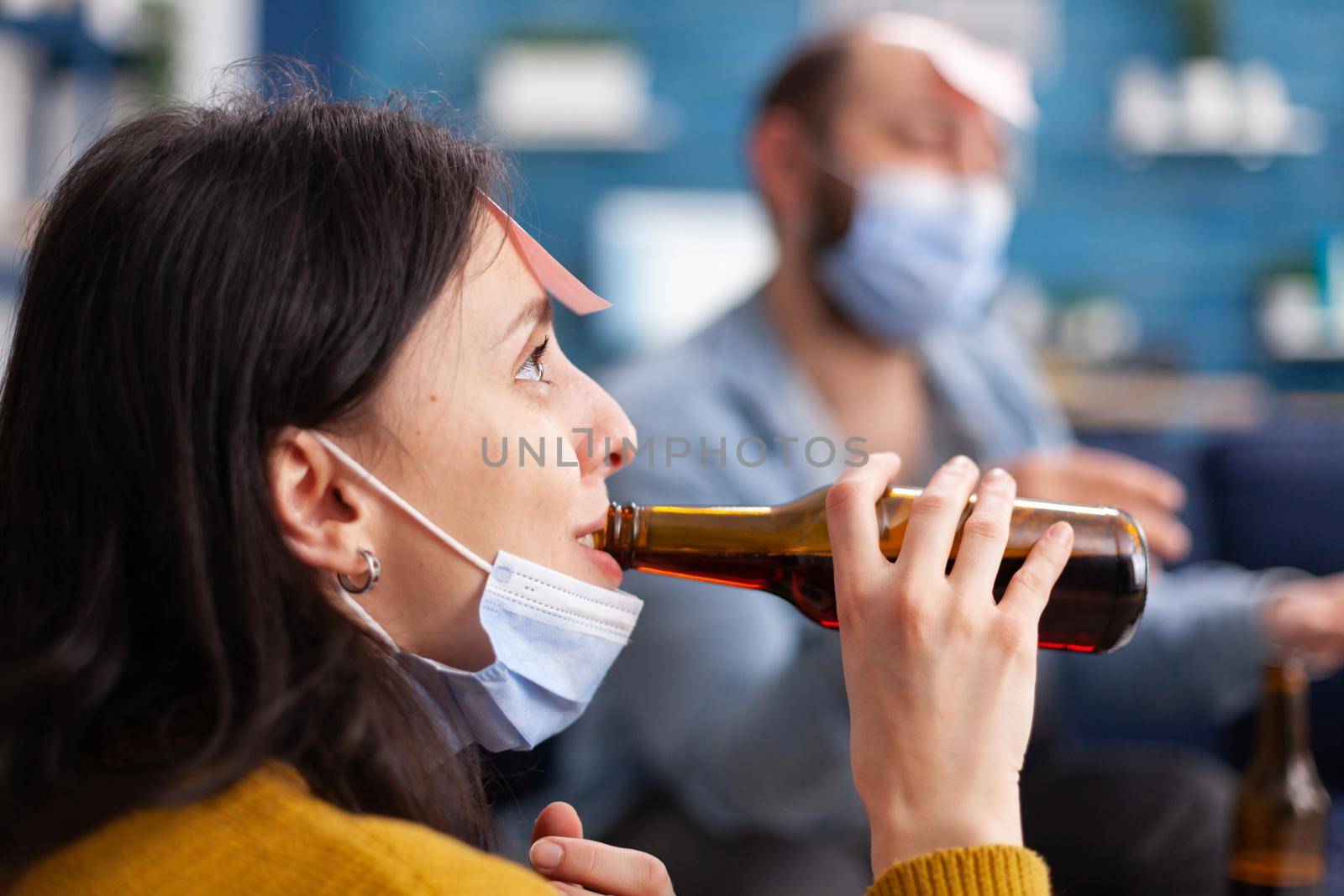 Close up of happy cheerful woman drinking beer while playing name game with multi ethnic friends having sticky notes on forehead wearing face mask keeping social distancing. Conceptual image.