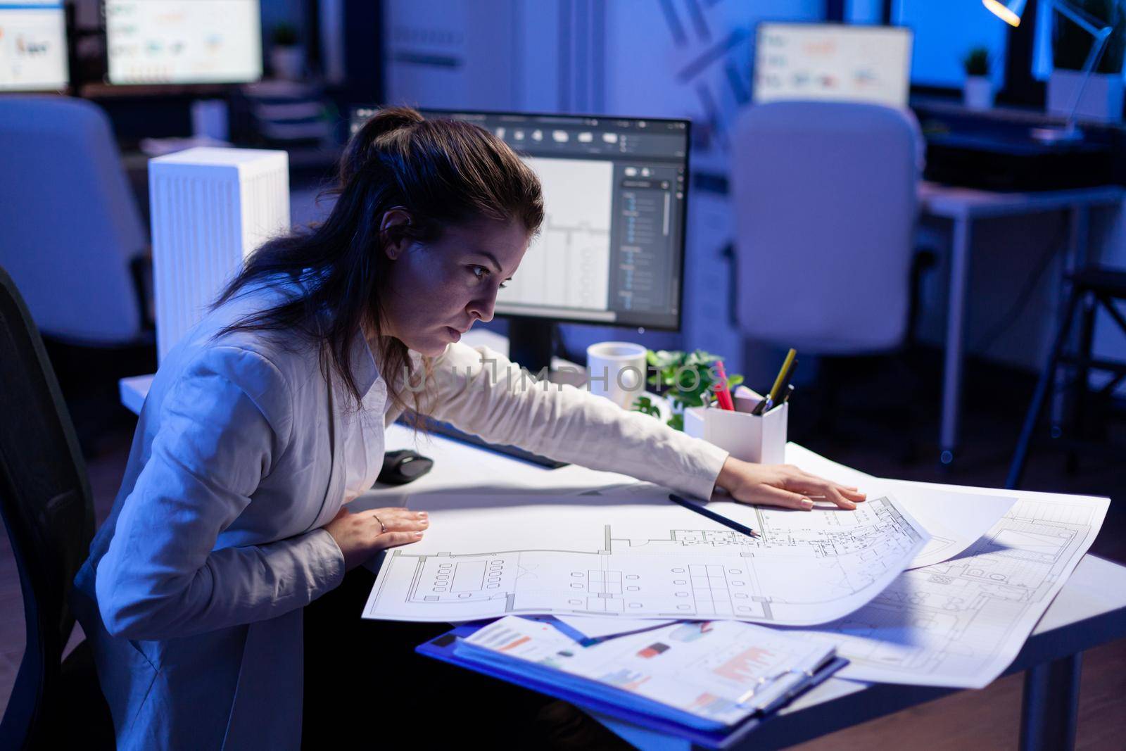 Overworked woman architect checking and matching blueprints sitting at office desk in front of computer. Perfectionist designer using arhitecture blueprints of buildings creating industrial prototype