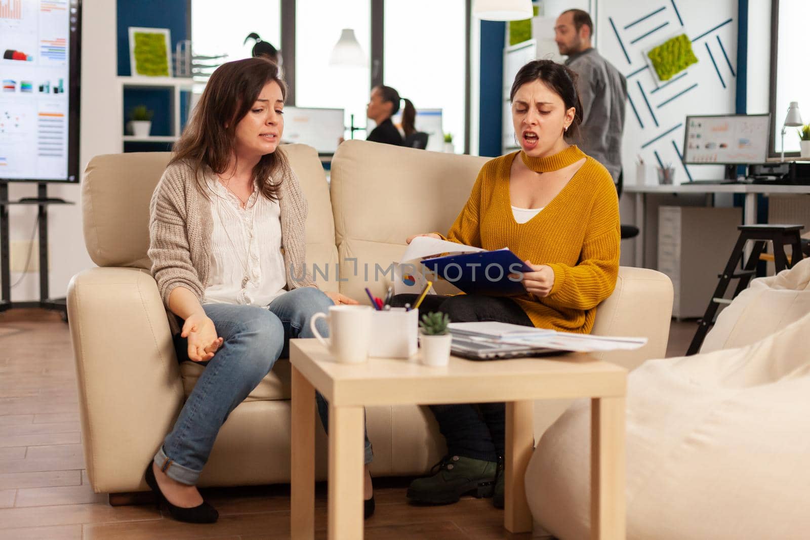 Woman leader yelling at employee sitting on couch in new start up business, upset of bad contract agreement. Women colleagues having big conflict, dispute about mistakes from project documents