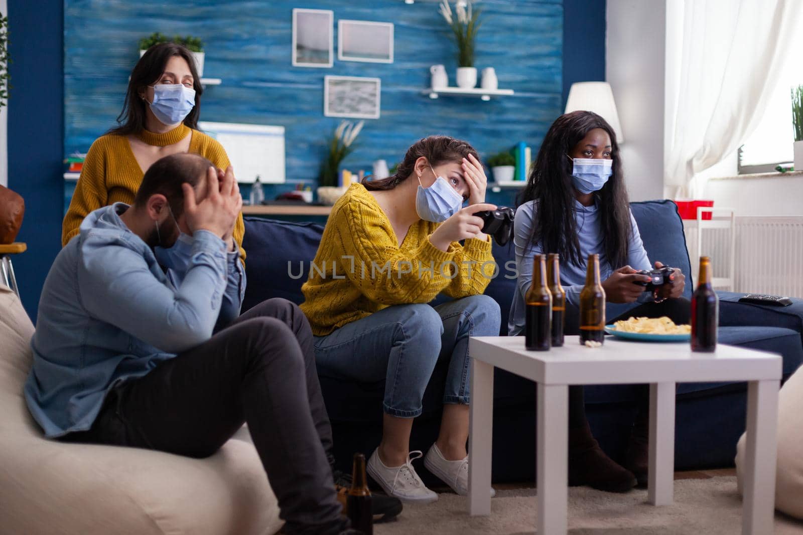 Group of mixed race friends losing while playing video games in home living room with joystick wearing face mask keeping social distancing in time of corona outbreak. Enoying beer and chips.