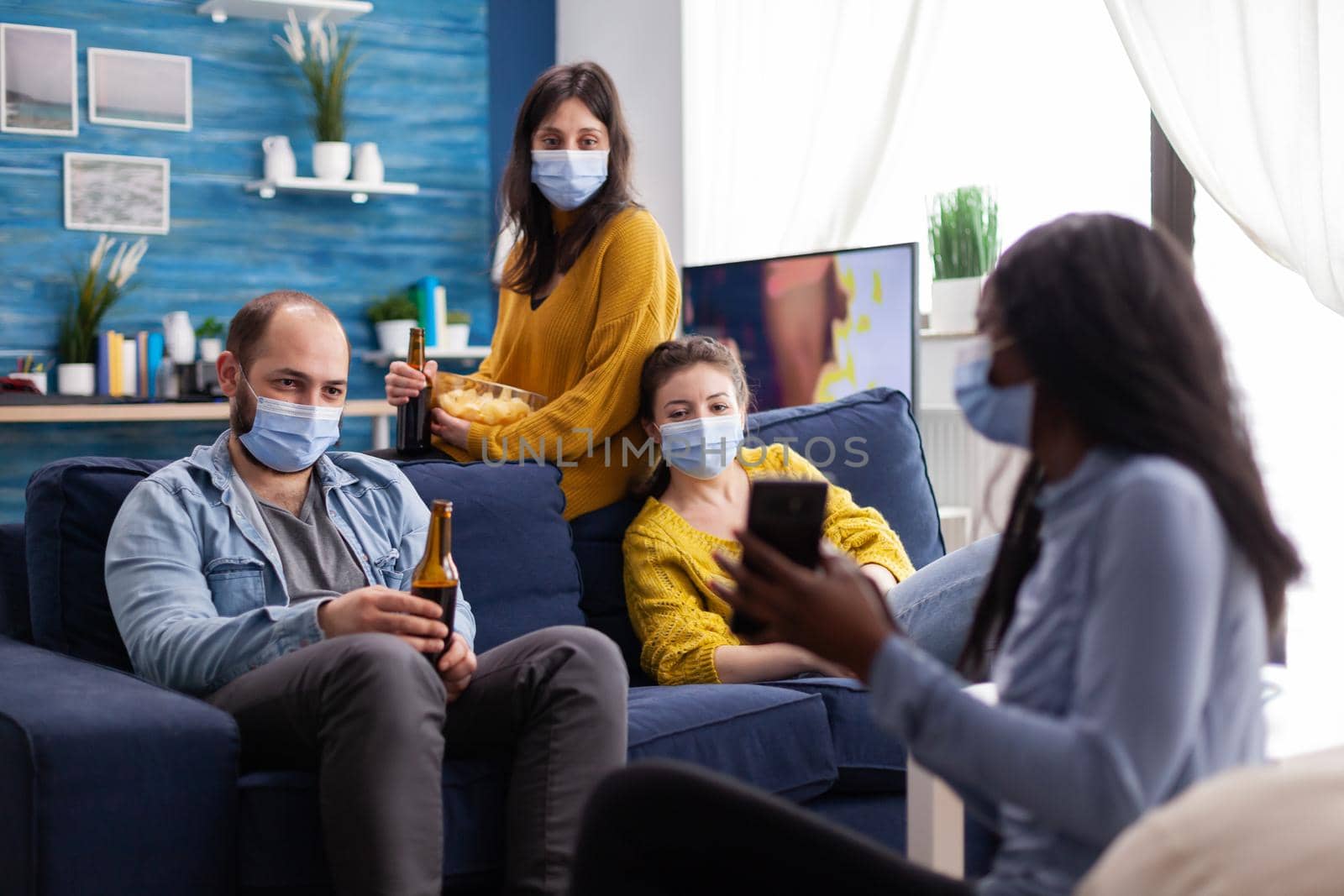 Mixed race cheerful friends looking at smartphone clip living room keeping social distancing to prevent coronavirus spread. Young man holding beer bottle. Conceptual image.