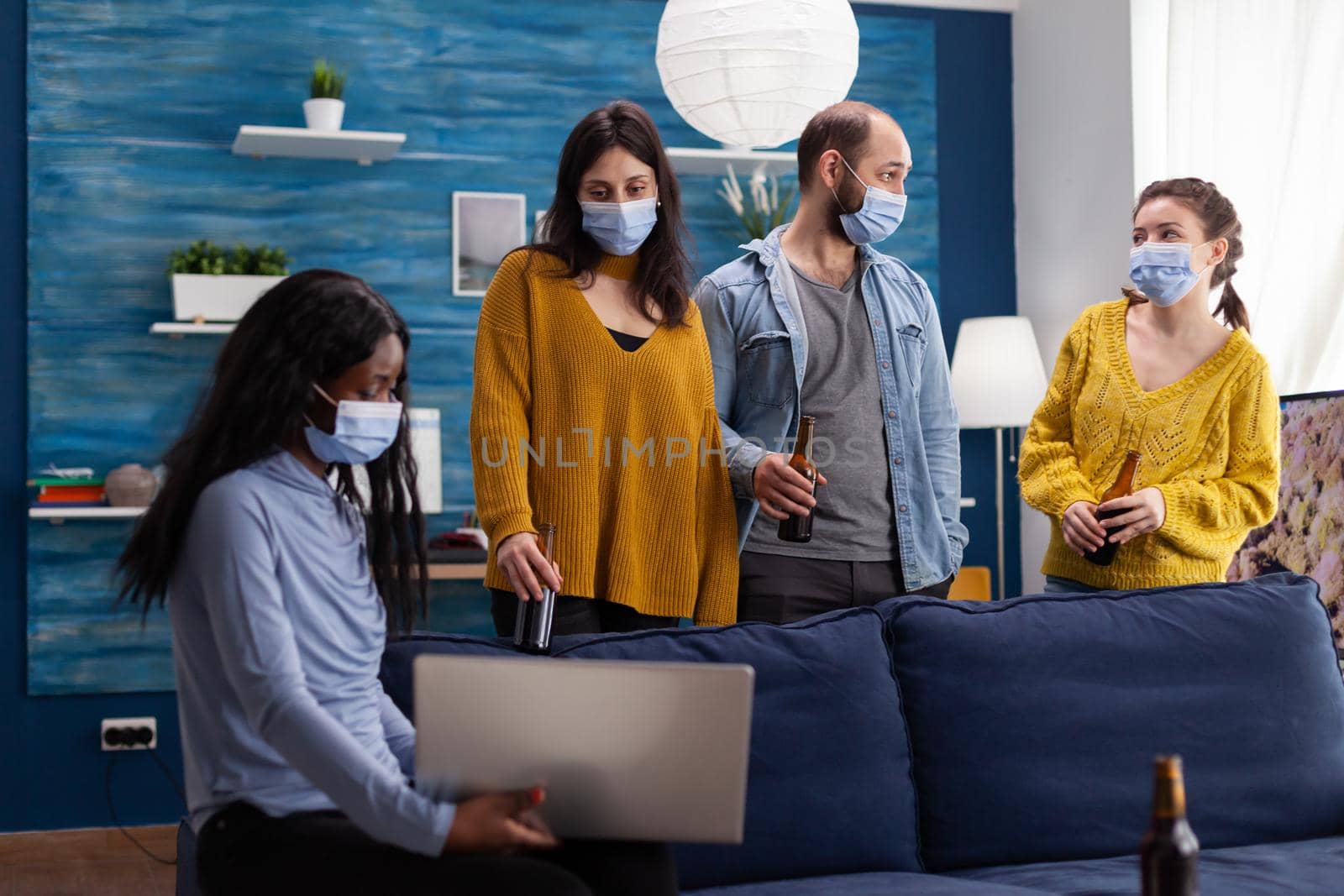 Multi ethnic friends wearing face mask drinking beer using laptop keeping social distancing to prevent coronavirus spread during global pandemic having fun in home living room. Conceptual image.