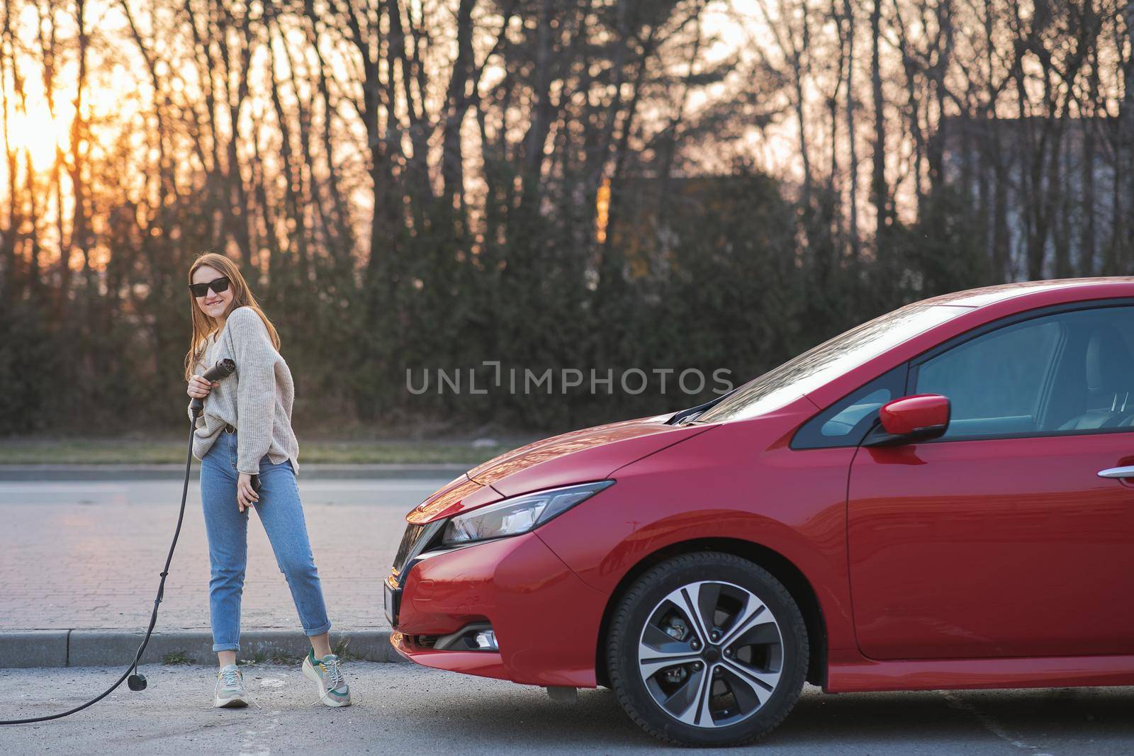 Young adult woman looking at camera having fun and holding in hand power cable supply standing near electric car at charging station