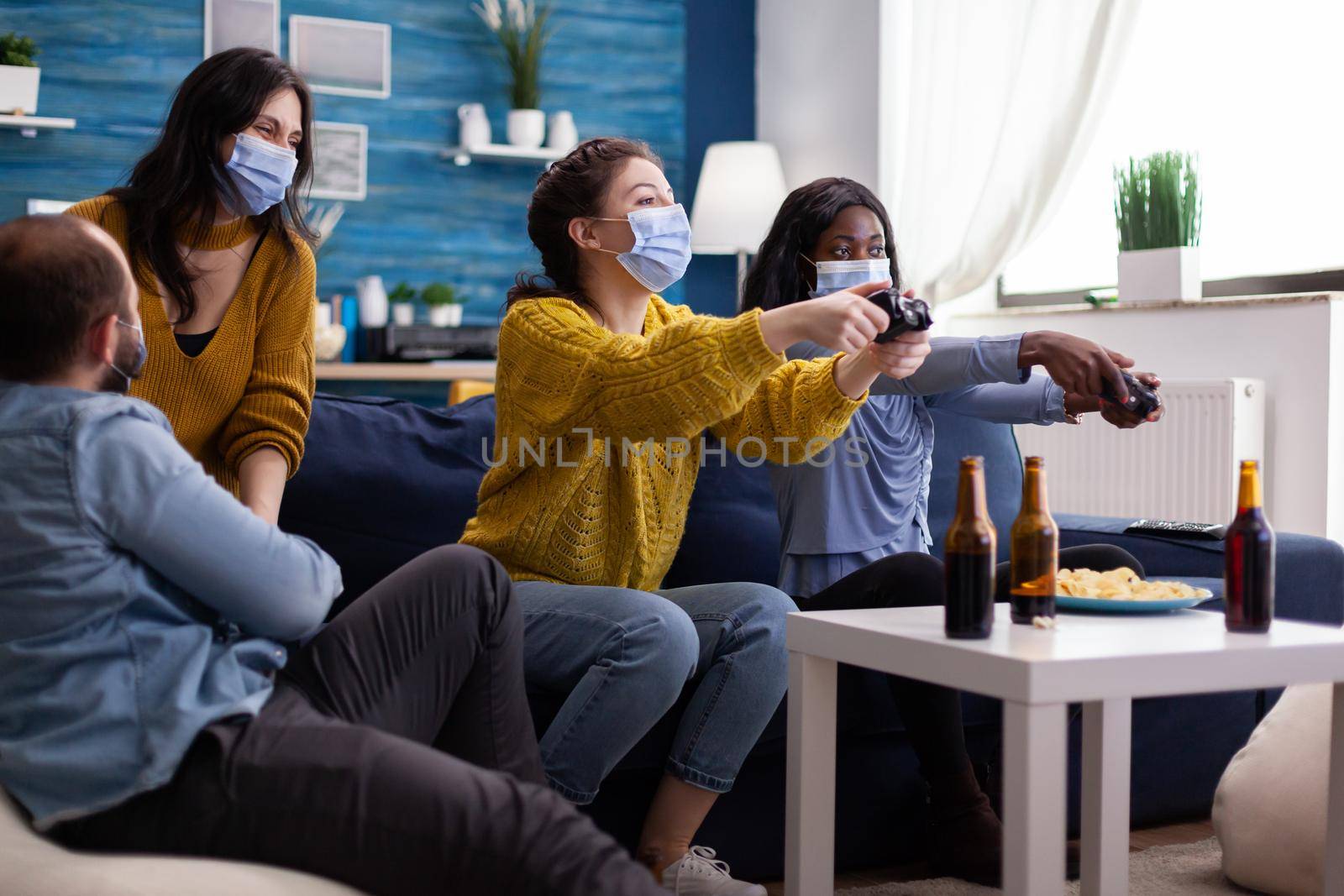 Mixed race friends meeting to playing video games with wireless controller wearing face mask to prevent getting sick with corona in time of social pandemic. Group of people enjoying time together.