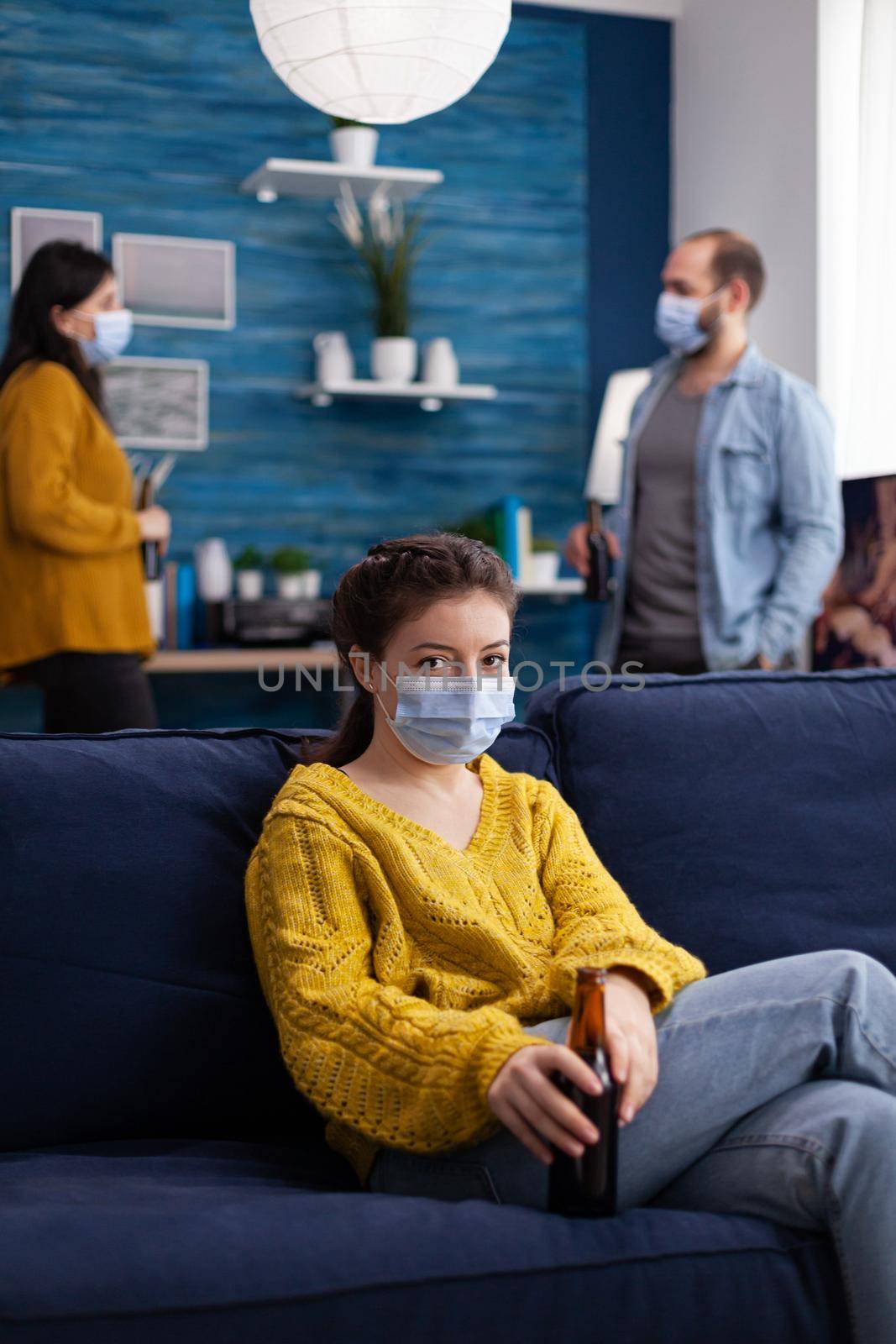 Happy young woman relaxing with friends sitting on couch wearing face mask as prevention against spreading coronavirus during global pandemic holding beer bottle looking at camera.