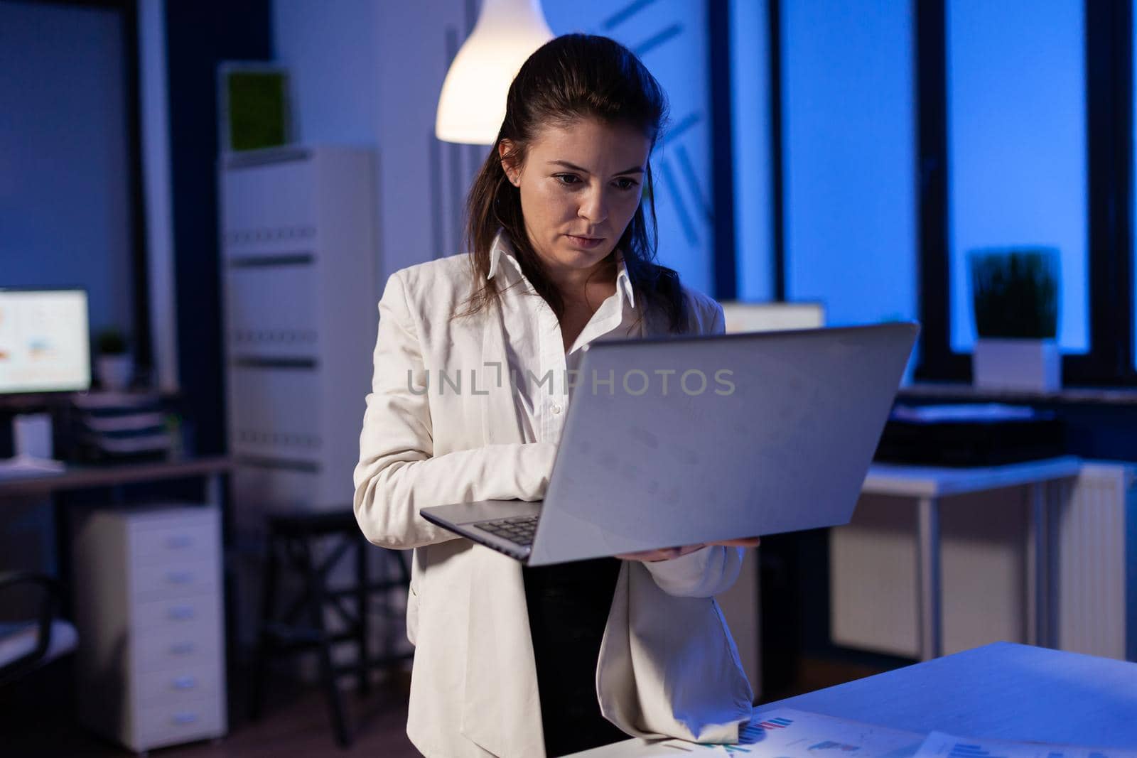 Tired entrepreneur analysing financial reports standing in start up office holding professional laptop. Focused employee calculate marketing profit late at night using modern technology network