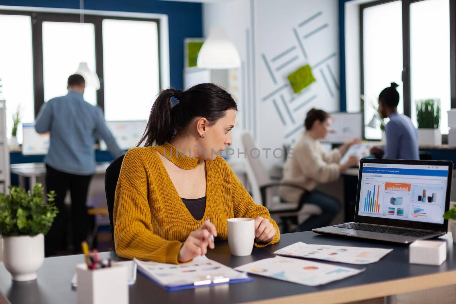 Businesswoman sitting at desk in office analyzing charts on laptop screen. Executive entrepreneur, manager leader sitting working on projects with diverse teamworkers.