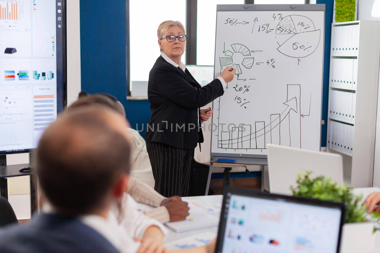Mature businesswoman writing on white board, presenting sales evolution answering question. Serious speaker boss executive, business trainer explaining development strategy to motivated mixed race employees.