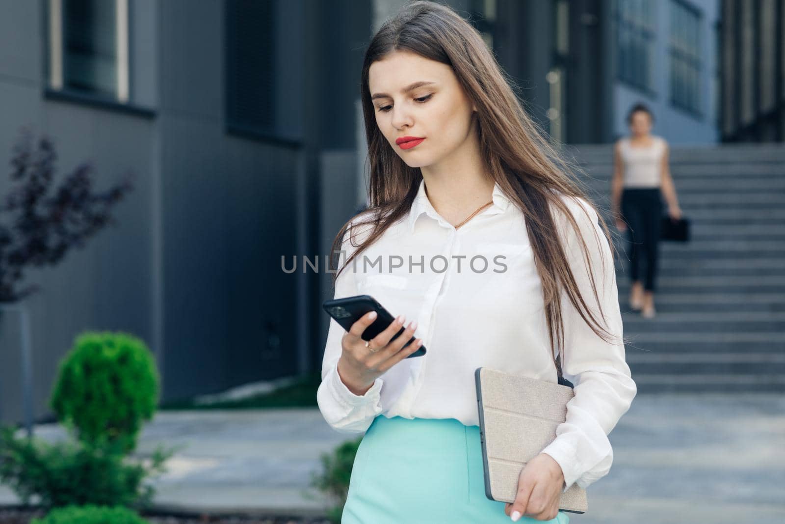 Stylish businesswoman using her phone outdoors. Beautiful female with long hair reading a message using her smartphone in front of building by uflypro