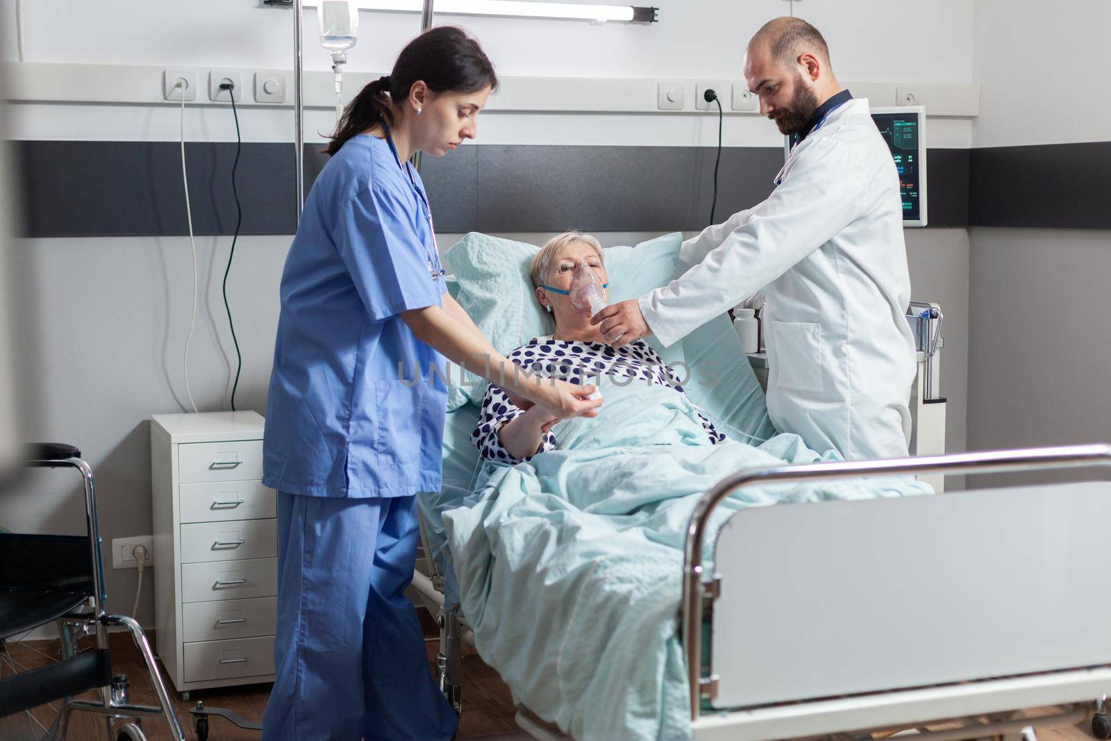 Hospitalized senior woman inhale and exhale through oxygen mask laying in hospital bed. Medical nurse using stethoscope listening patient heart.
